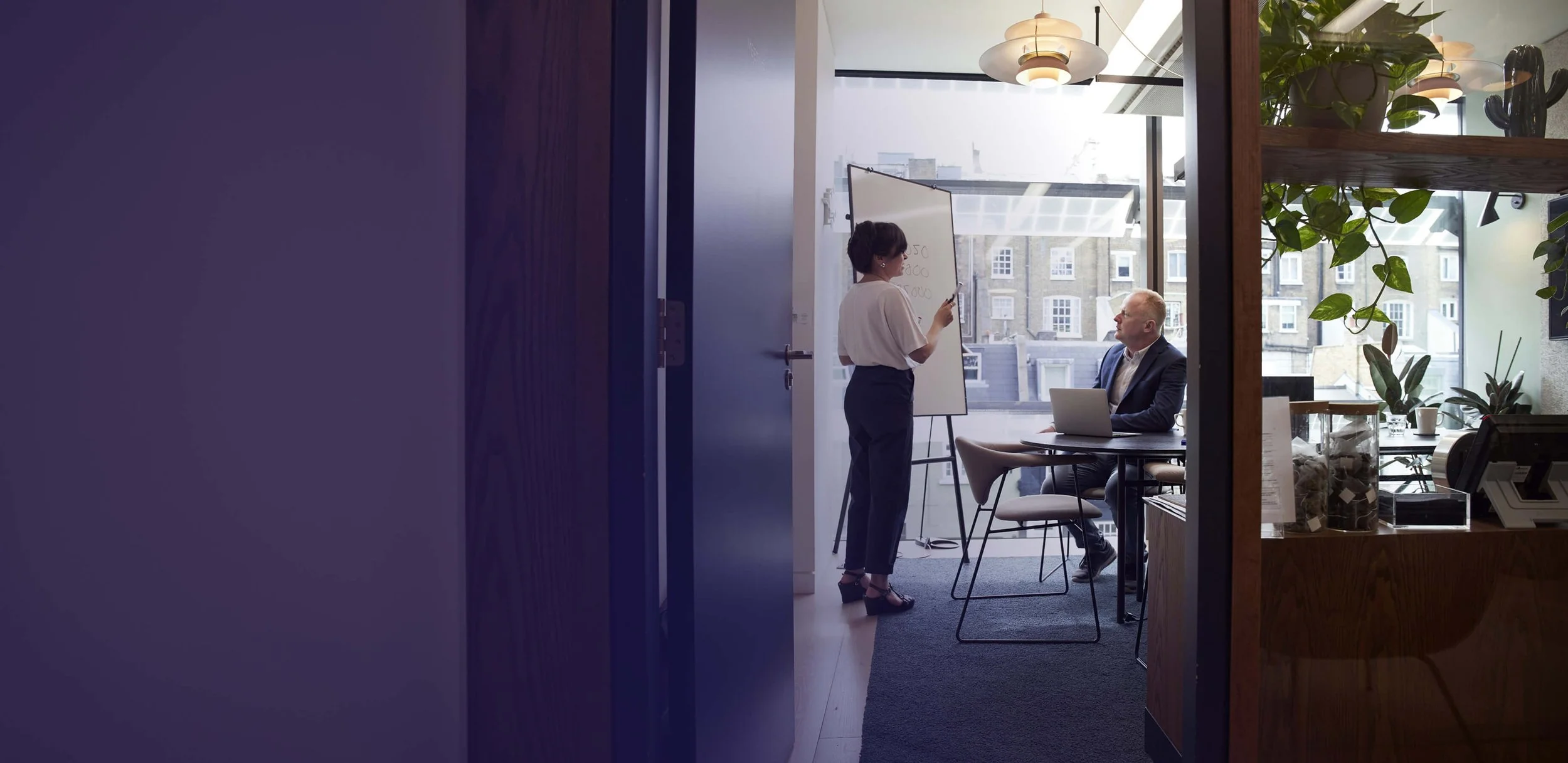 A woman presents on a whiteboard to a colleague with a laptop in a modern, purple-accented office with city views.