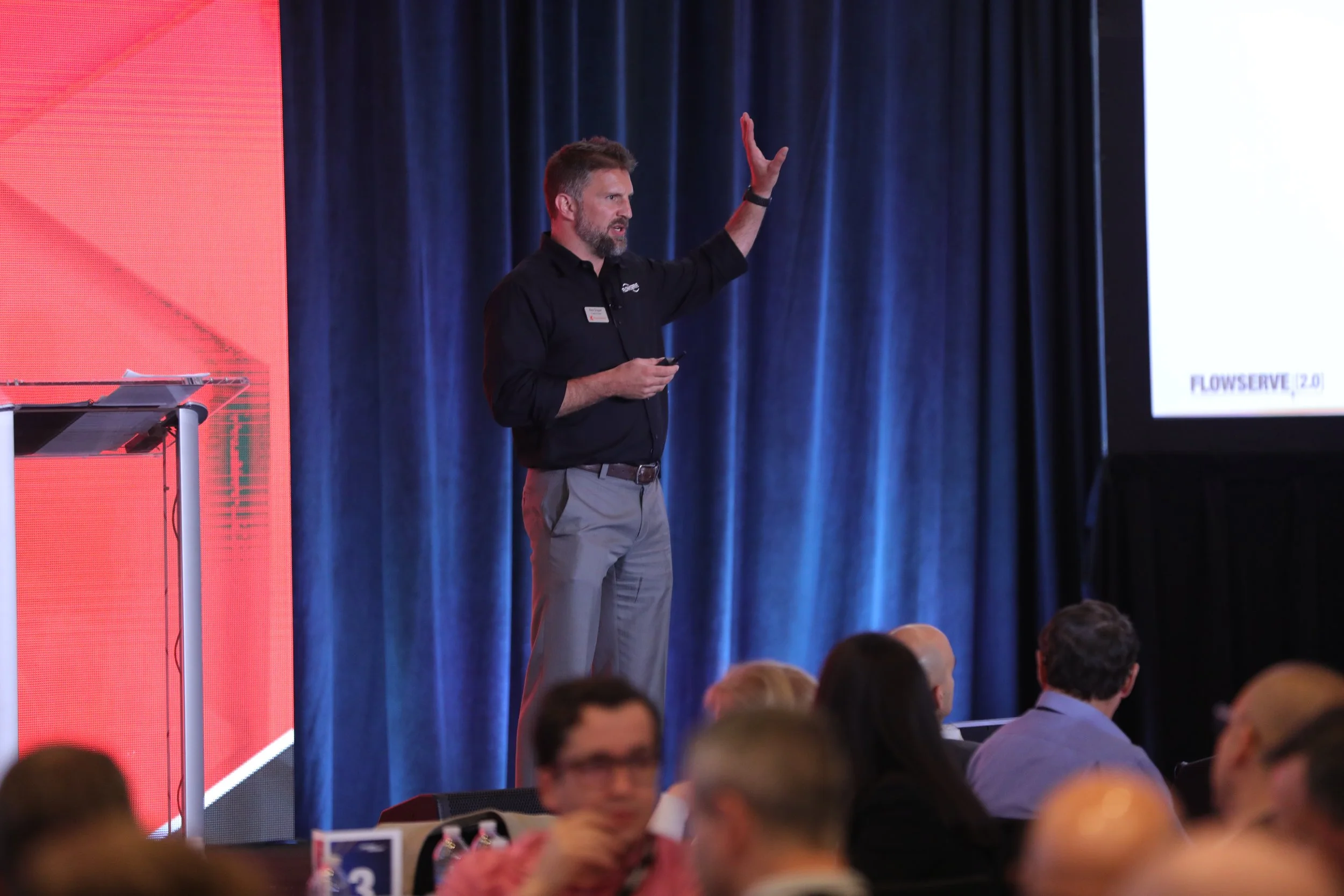 A man giving a presentation on a stage, standing with one hand raised and holding a clicker or phone in the other, in front of an audience with many people seated at tables.