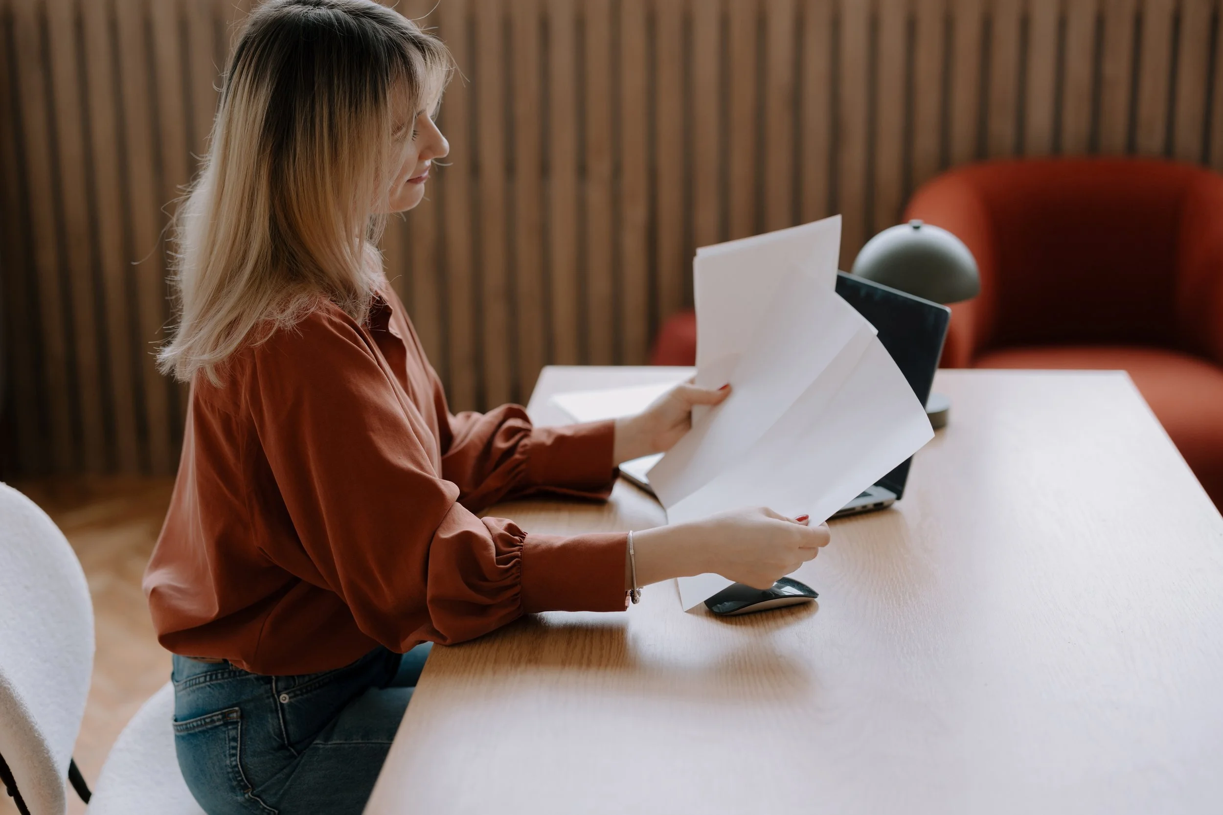 A woman with blonde hair sitting at a wooden desk, reviewing papers, with a laptop and a green desk lamp on the desk, and a red cushioned chair behind her.