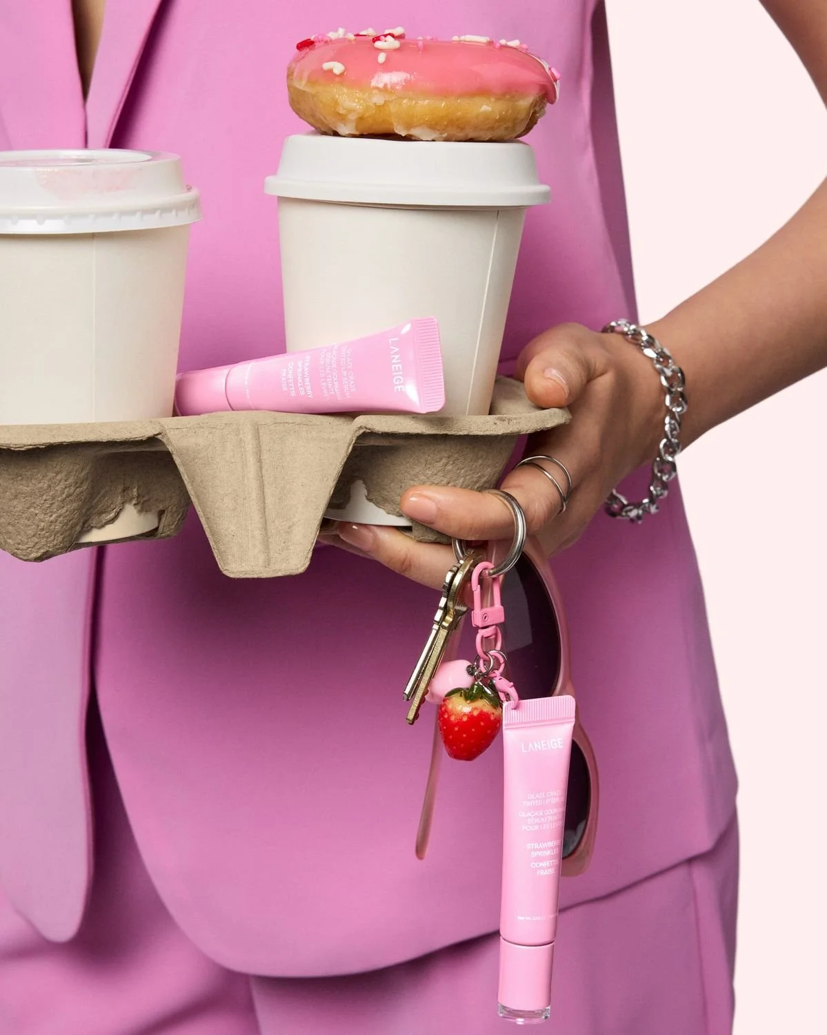 A person holding a tray with two white paper coffee cups, a pink glazed donut with sprinkles on top, and a pink skincare tube. The person has a silver bracelet, rings, and a keychain with a strawberry charm and sunglasses attached, all against a pink background.