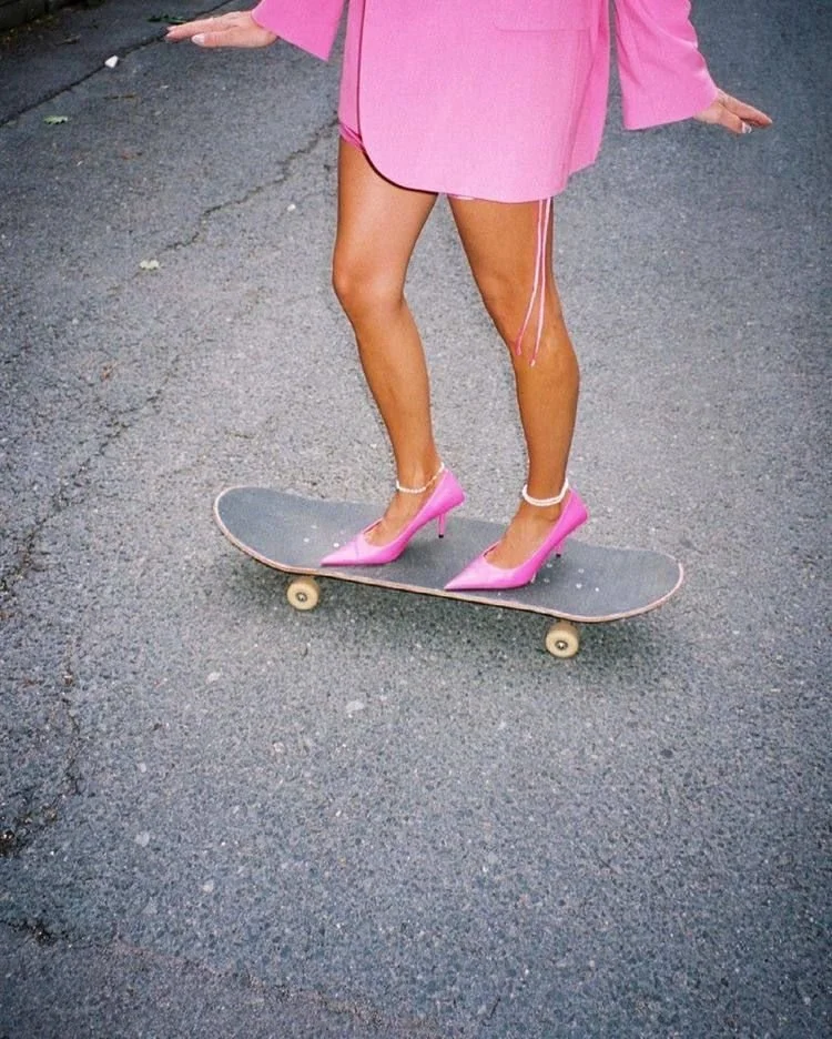 A person wearing pink high heel shoes and a pink dress is standing on a skateboard on an asphalt road.