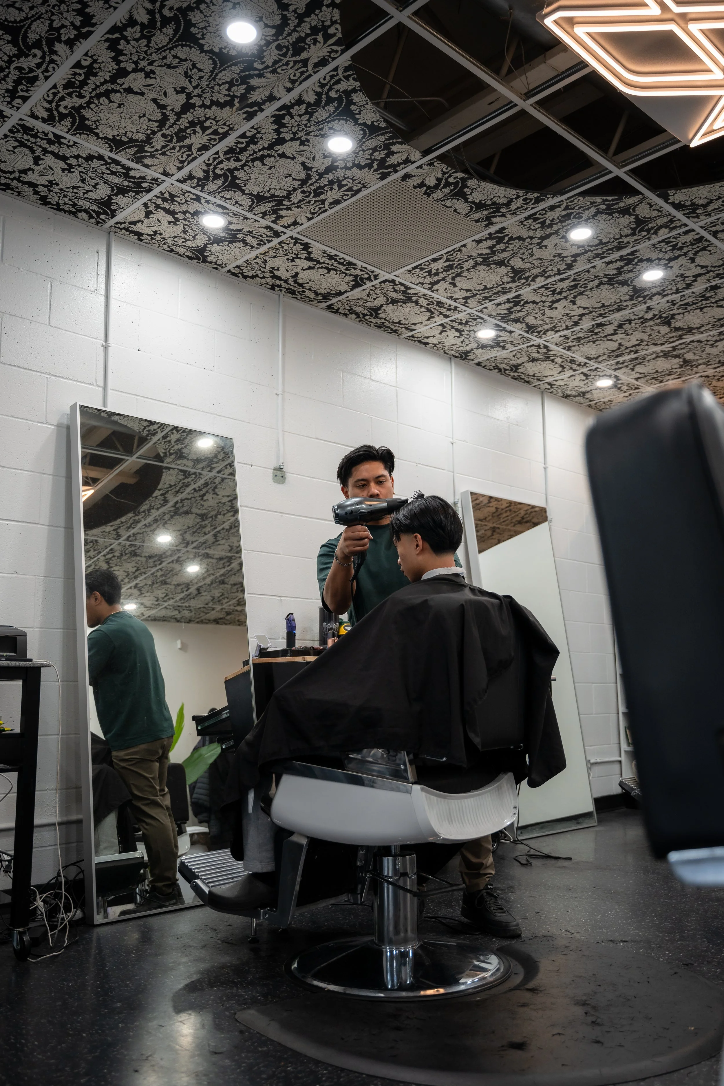 Barber blow-drying a customer's hair in a modern hair salon with patterned ceiling tiles and large mirrors.