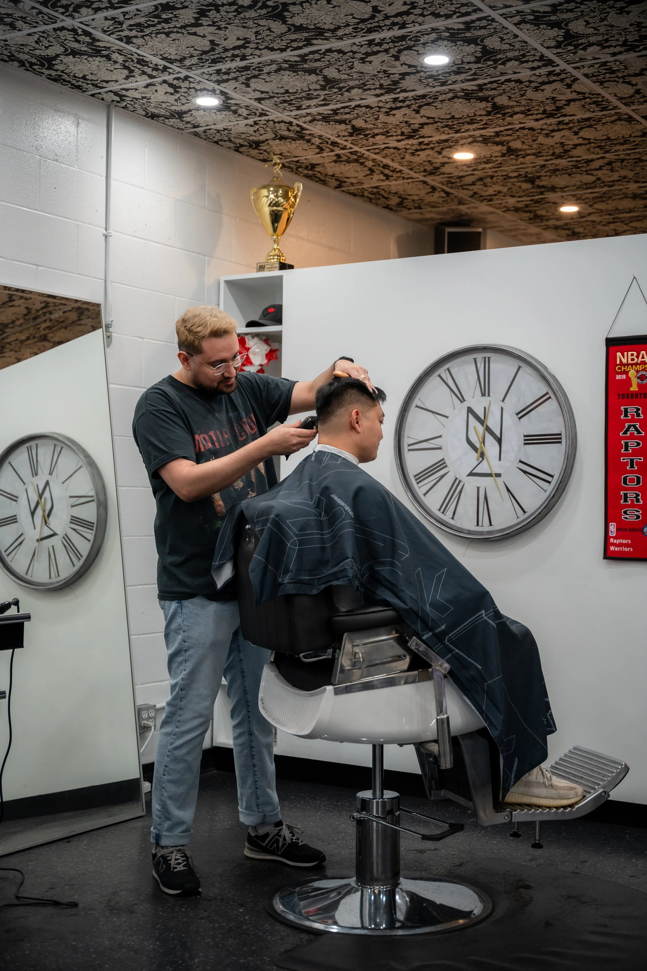 A barber giving a haircut to a man sitting in a barber chair inside a salon, with clocks and a sports banner on the wall.