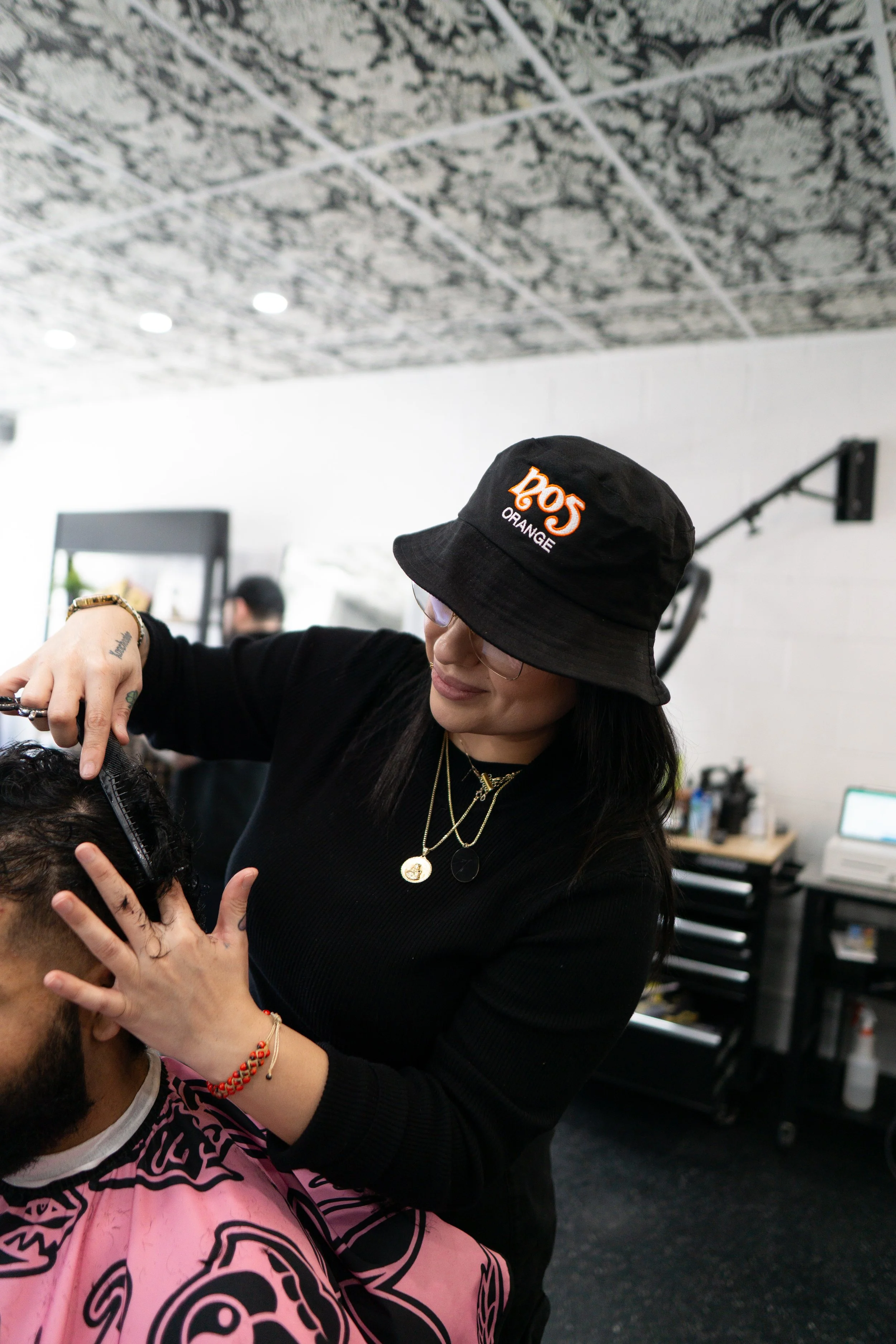 A woman wearing a black bucket hat with orange and white text, black top, and gold jewelry is cutting a man's hair in a salon.