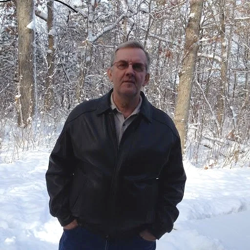 A man standing outdoors in a snowy forest, wearing a black jacket and glasses.