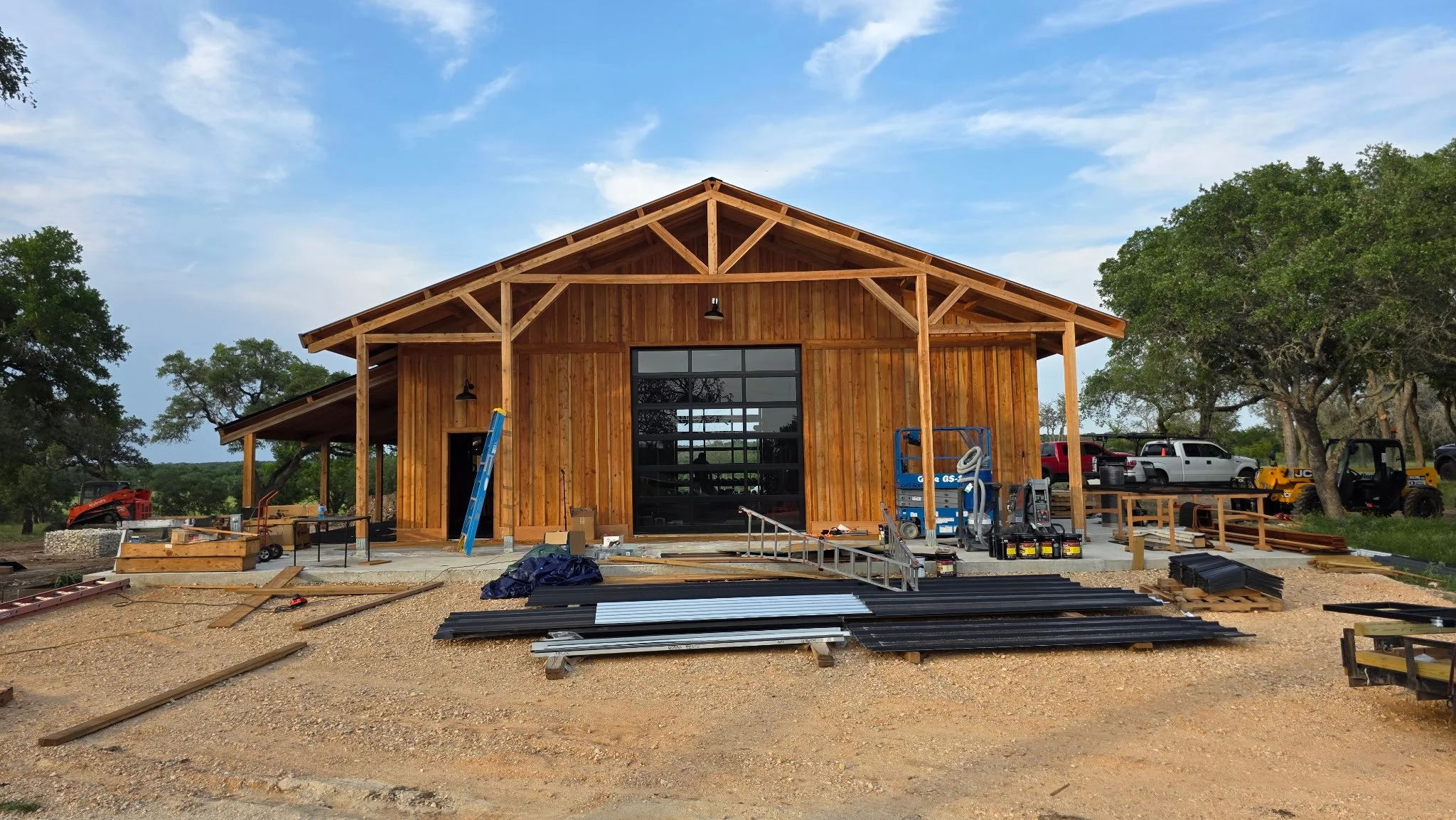 A wooden building under construction with a large glass garage door in the center, surrounded by construction equipment and materials, with trees and parked trucks in the background.