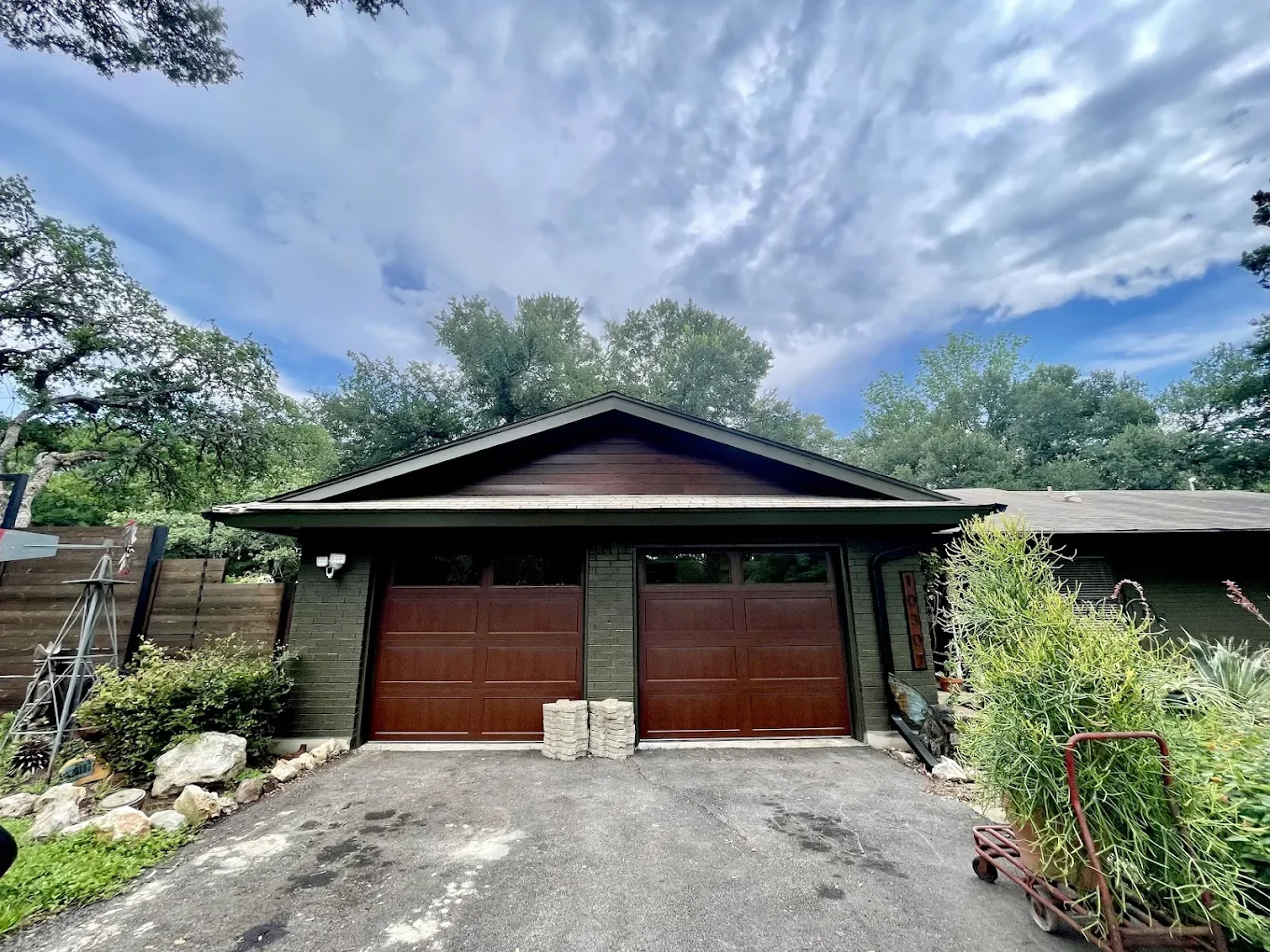 Front view of a house with a double garage door, surrounded by trees and plants, with a cloudy sky overhead.