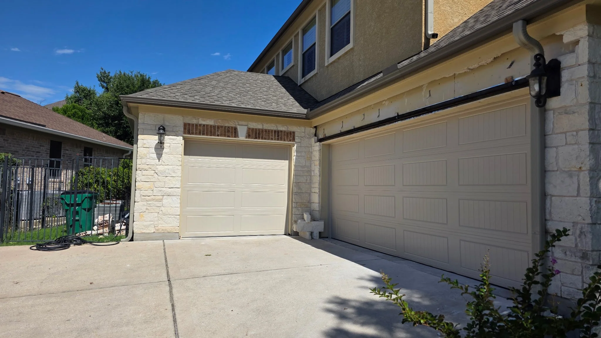 A residential house with two beige garage doors, stone accents, porch light, and a driveway with a garden hose on the ground, under a clear blue sky.
