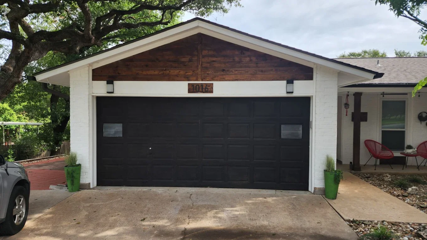 front view of a house with a black garage door, white brick walls, and a small porch with two red chairs. A large tree is on the left, and two green plant pots are flanking the driveway.