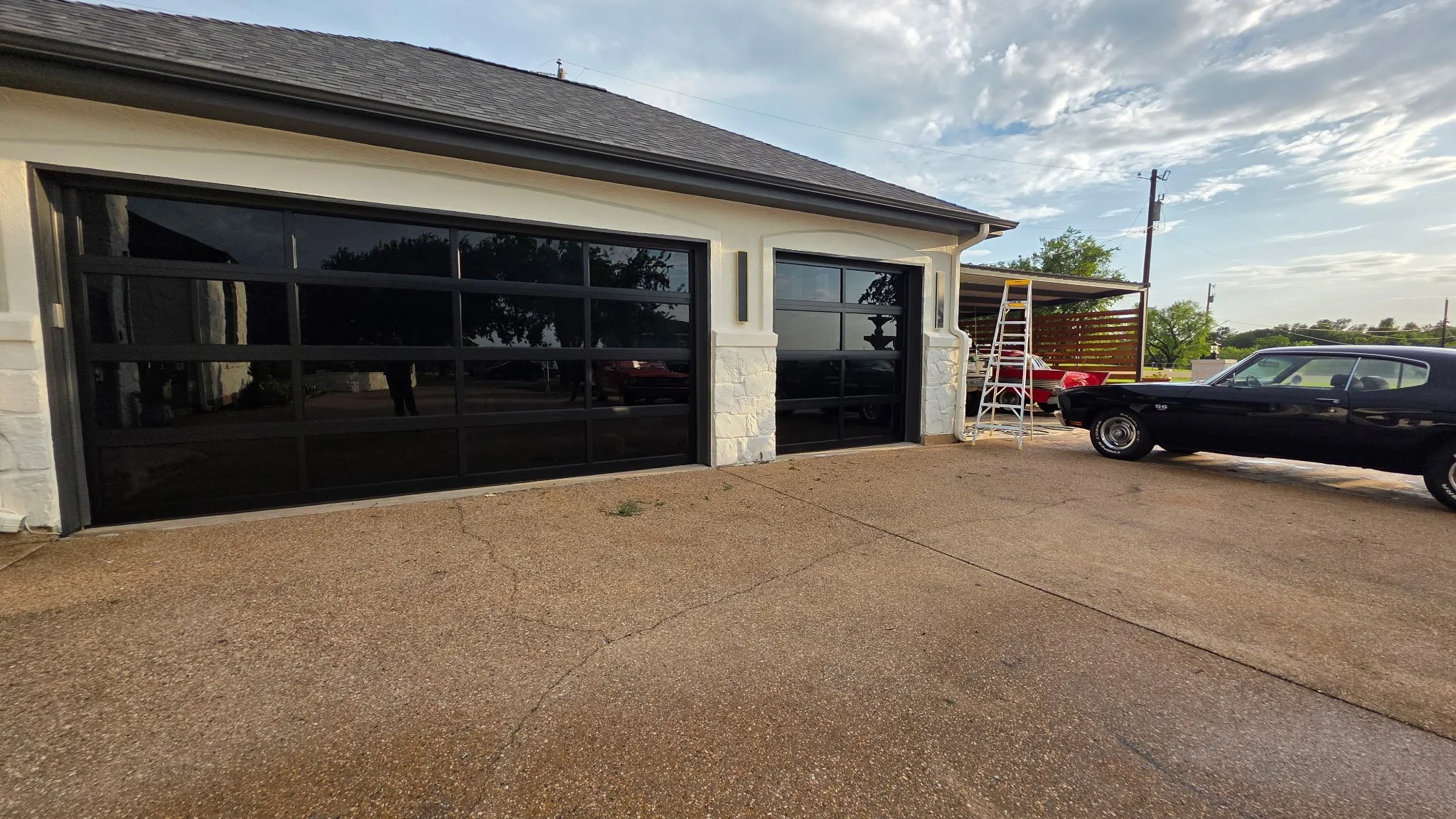 A driveway in front of a garage with large glass doors. There are two vintage cars parked on the right side, one black and one red. A ladder and a wooden slatted privacy screen are seen in the background. The sky is partly cloudy.