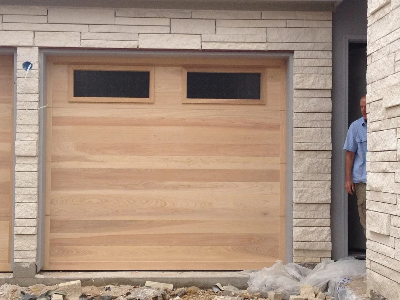 A newly installed wooden garage door with a two-pane window at the top, surrounded by a brick exterior wall, with a man standing in a doorway to the side.