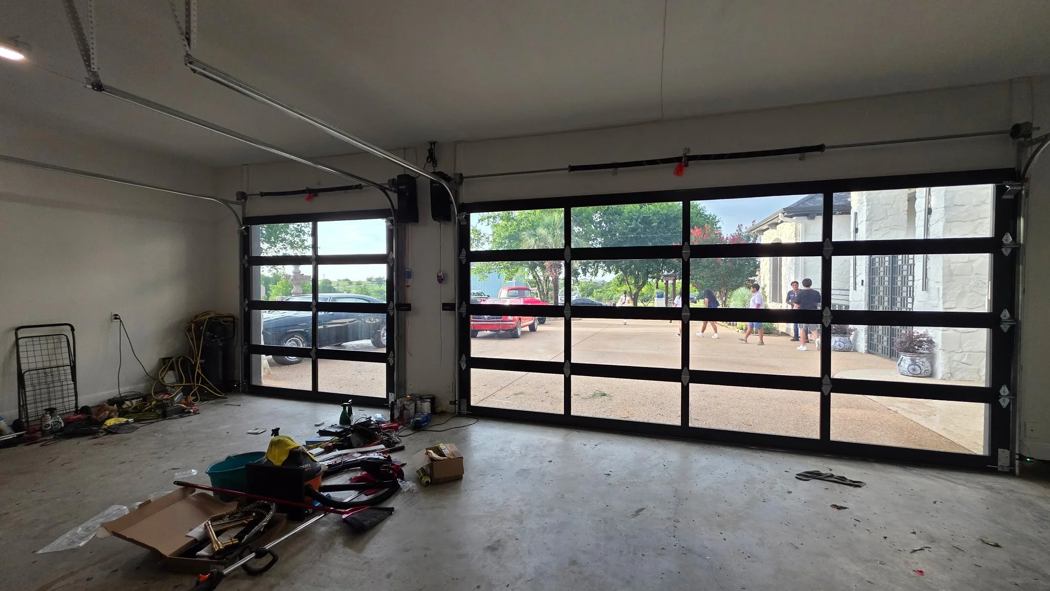 An unfinished garage interior with large black-framed glass garage doors, some tools and equipment on the floor, and a view of parked cars and people outside.