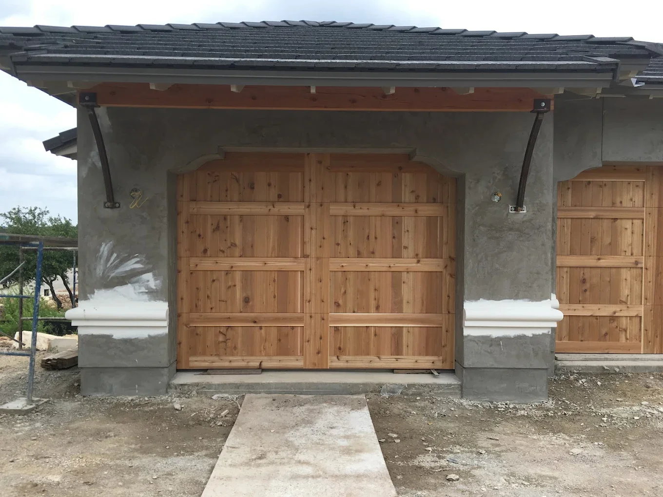 Wooden garage doors installed on a concrete and stucco structure with unfinished landscaping in foreground.