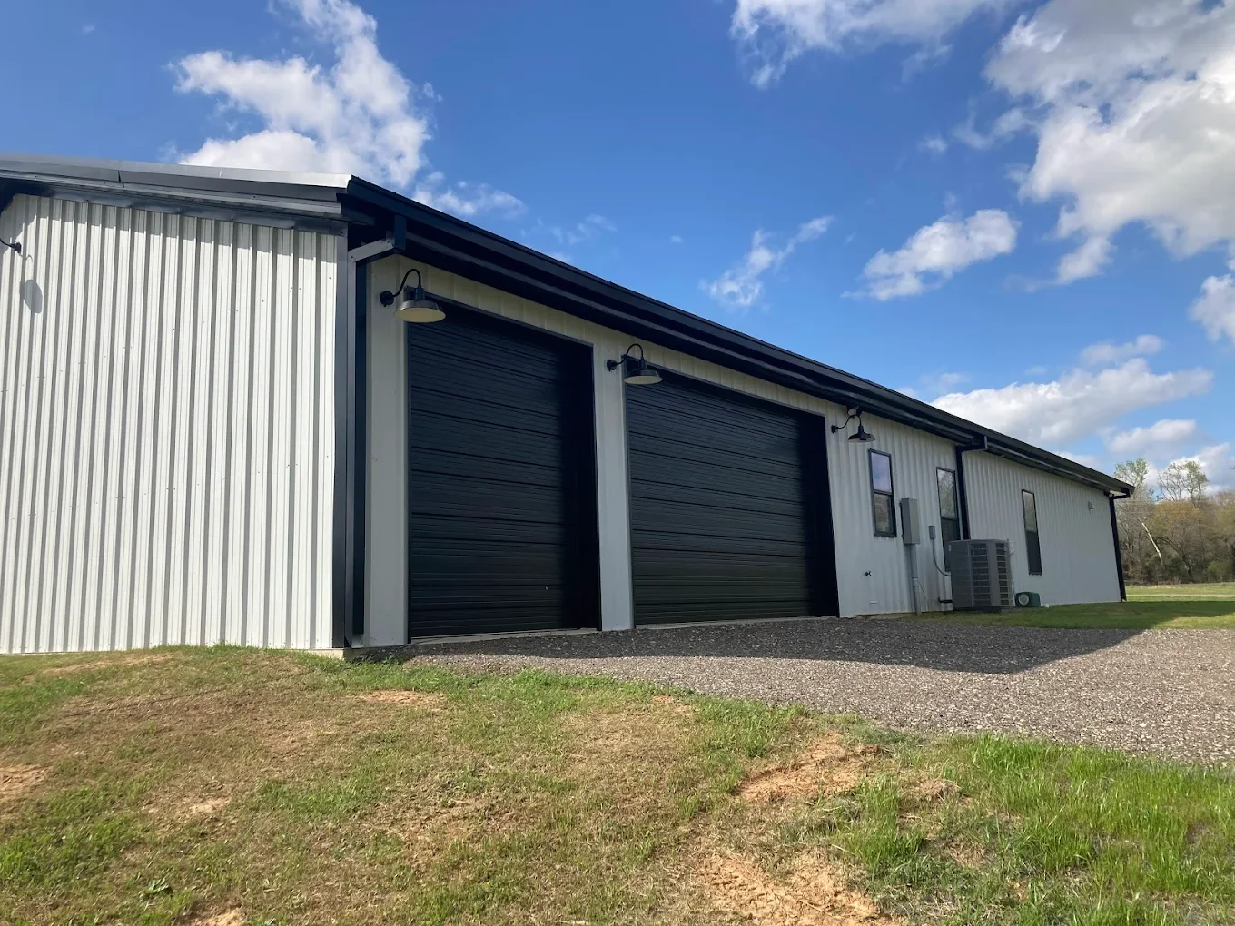 Metal building with two large black garage doors, black exterior lights, windows, and an HVAC unit outside on a sunny day with blue sky and scattered clouds.