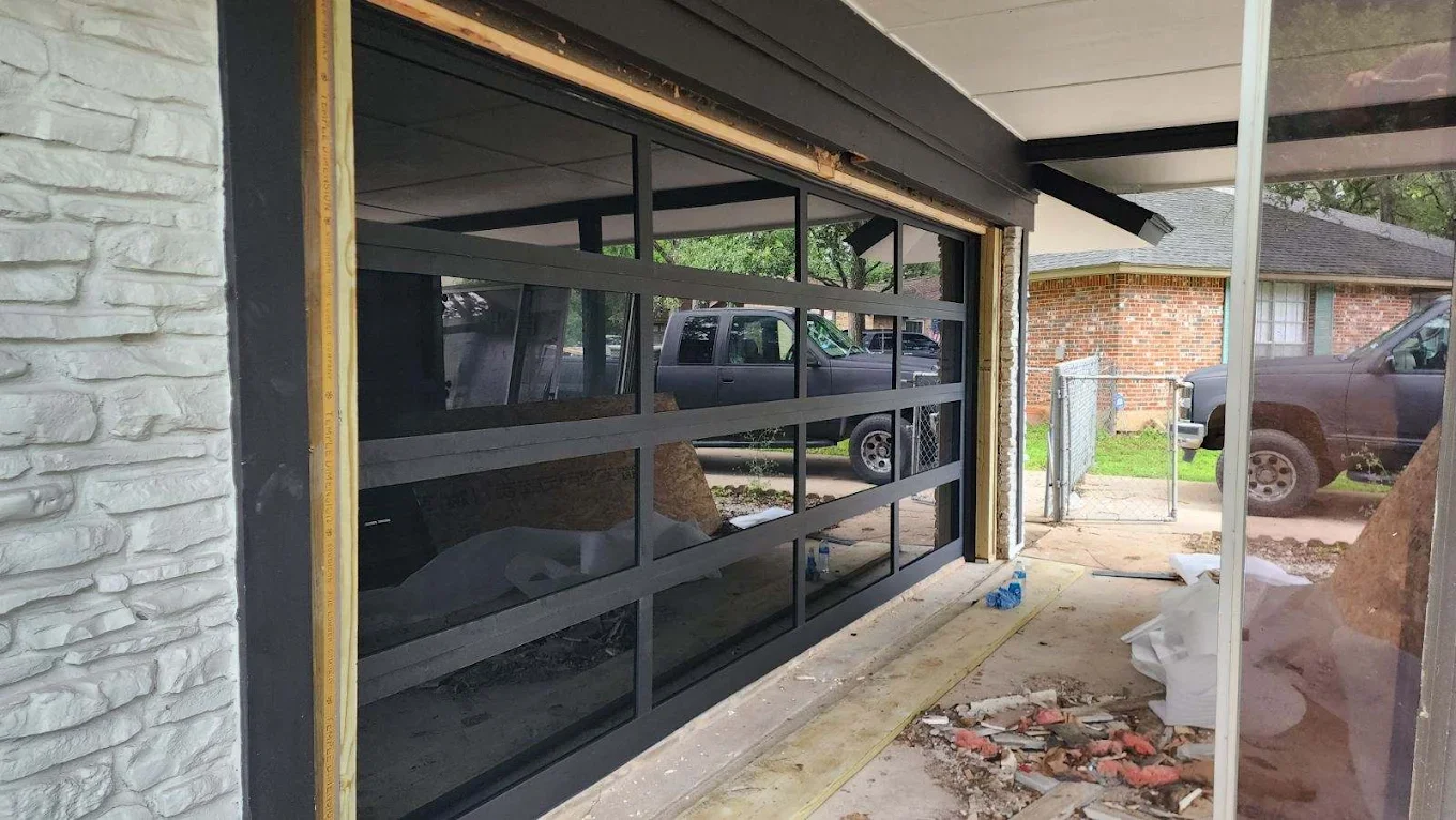 A garage under construction with a black-framed glass garage door. Construction materials and debris are present on the ground inside and outside the garage. There is a white brick wall on the left and a small outdoor patch with a chain-link fence an