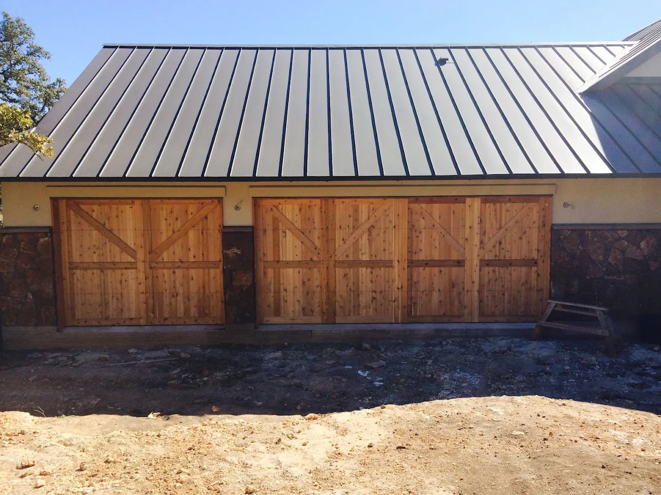A building with a metal roof and two wooden doors, with a stone wall section and a small wooden bench nearby.