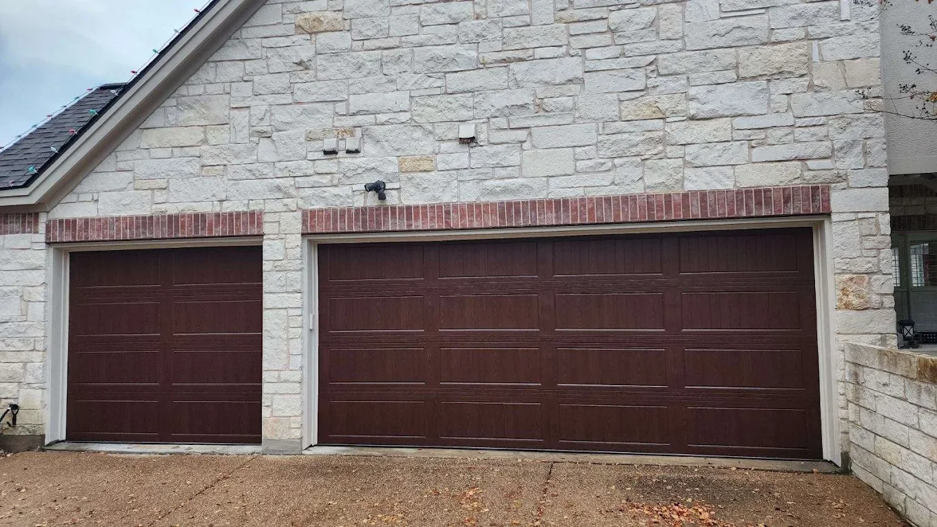Two closed garage doors made of dark wood in a stone house, with a small black security camera above the doors and a gravel driveway in front.