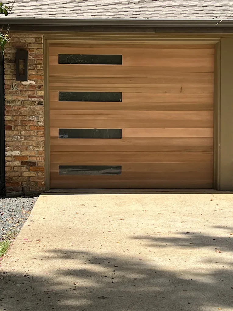 A modern wooden garage door with horizontal panels and rectangular glass inserts, adjacent to a brick wall and concrete driveway.