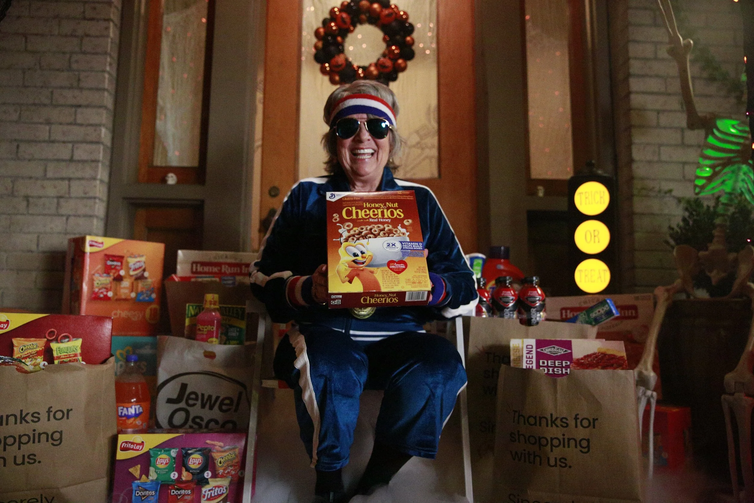 Woman dressed in 70s style clothing, wearing sunglasses, a headband, and a blue velour tracksuit, sitting on a chair outside a house at night, surrounded by grocery bags filled with snacks and beverages, including cereal boxes and soda bottles, smili
