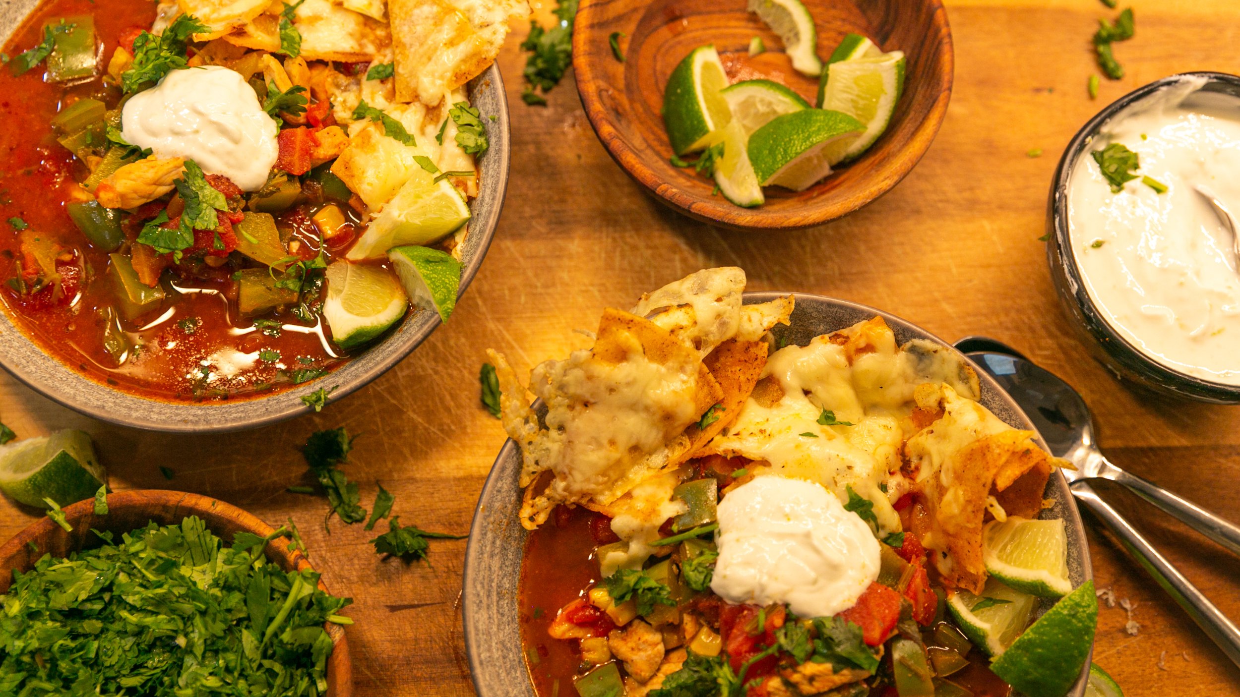 Two bowls of tortilla soup topped with cheese and sour cream, surrounded by lime wedges, cilantro, a bowl of salsa, and a bowl of sour cream on a wooden table.
