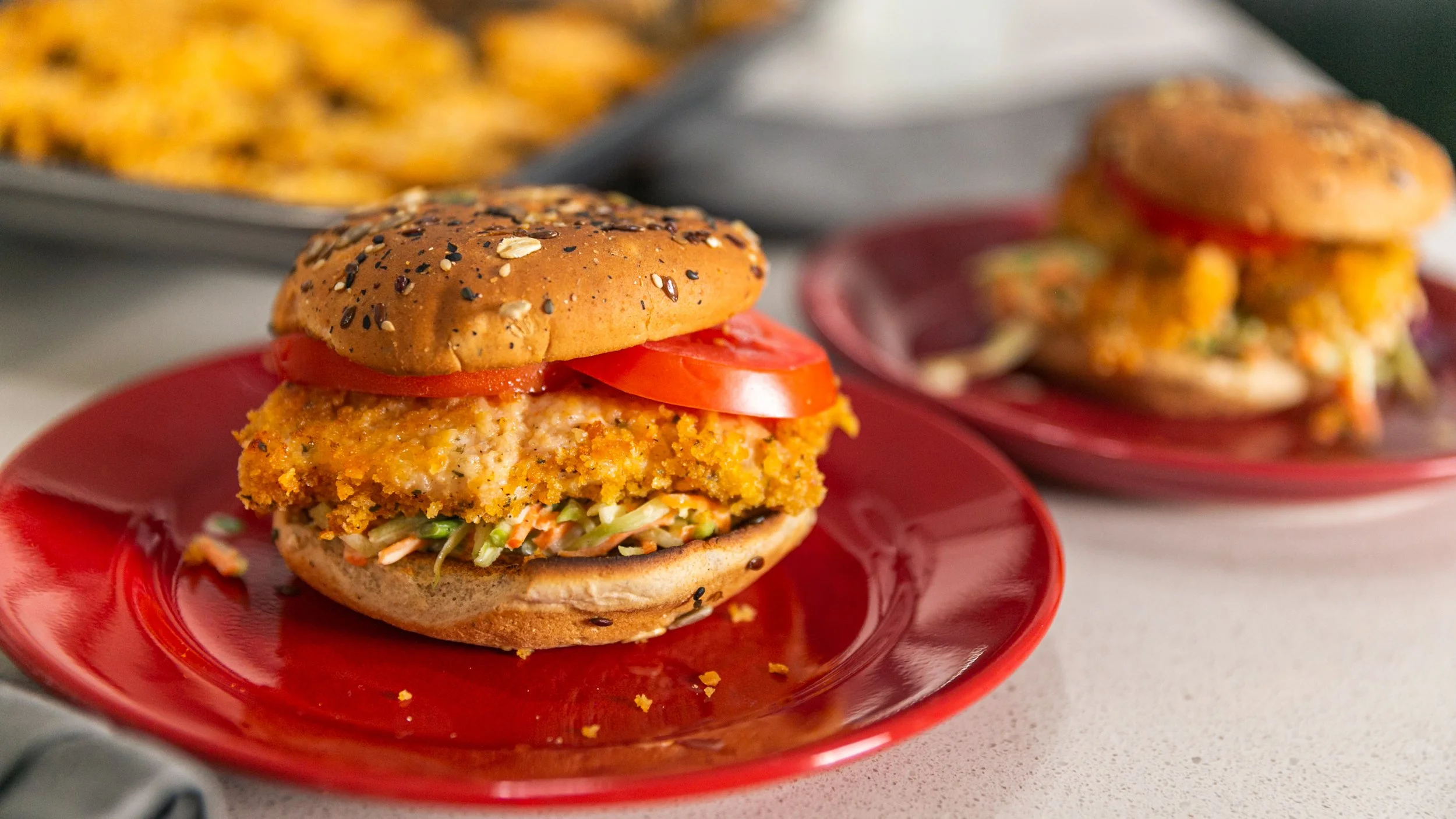 A veggie burger on a red plate with tomato slices and coleslaw underneath the patty, with another similar burger in the background.