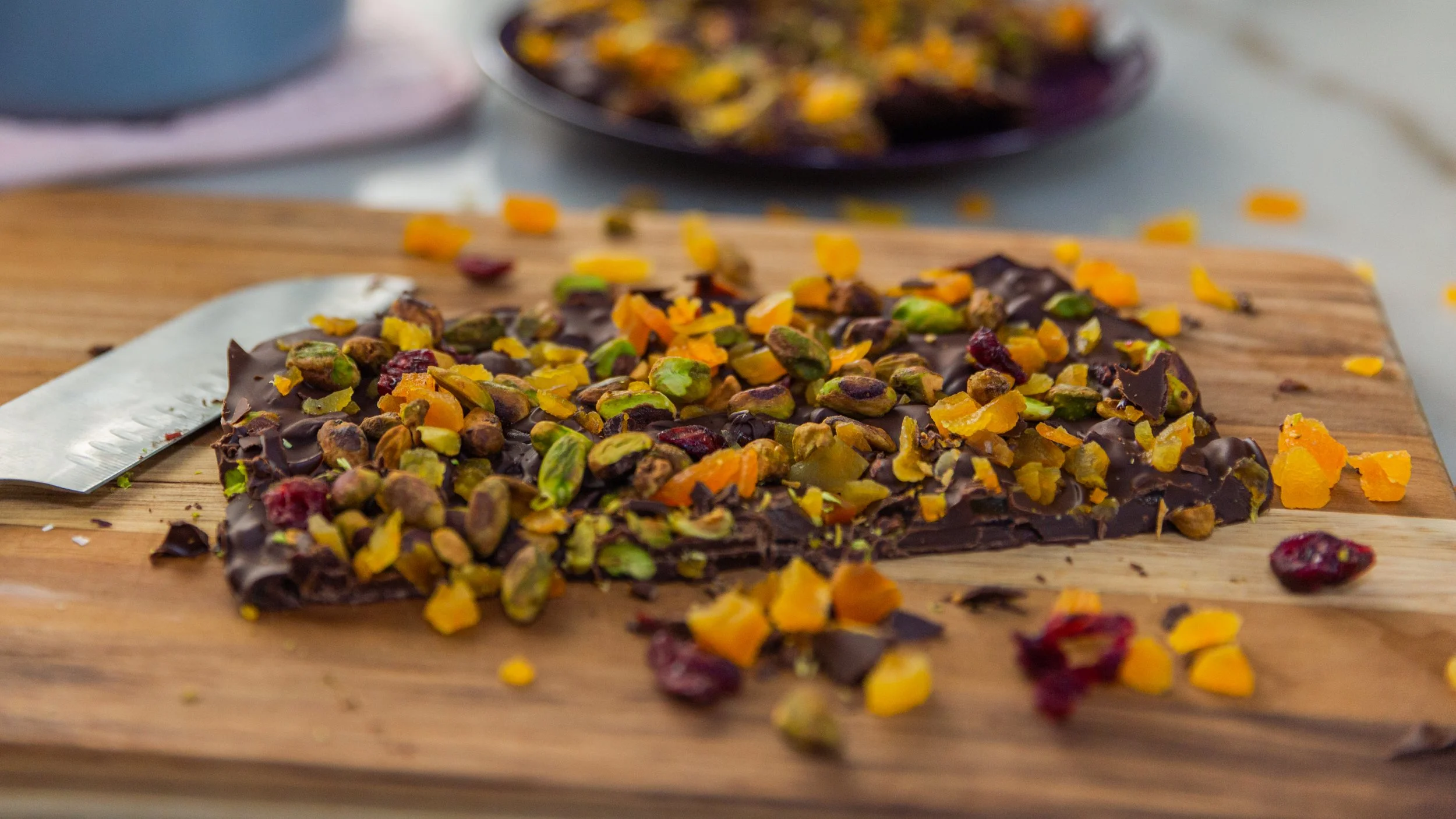 A piece of dark chocolate bark topped with chopped pistachios, dried apricots, and dried cranberries on a wooden cutting board.