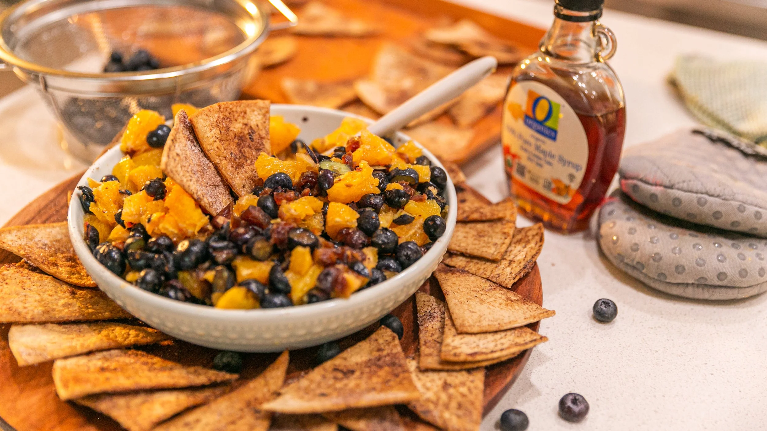 A bowl of mixed fruit salad with chopped mango, blueberries, and chopped peaches, tsurrounded by toasted pita chips, with a bottle of honey syrup and oven mitts nearby on a kitchen counter.