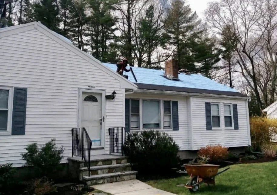 Person on roof installing or repairing a blue roof on a white house with black shutters, front yard with bushes, a wheelbarrow, and a sidewalk.