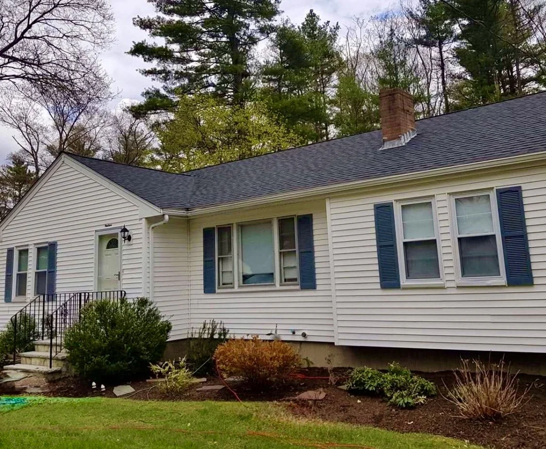 A white house with blue shutters, a black front door with a small overhang, and a brick chimney. There's a small set of steps with a black railing leading to the front entrance, surrounded by shrubs and bushes. The house has a gutter system and is situated in a yard with grass and landscaped garden beds. Tall trees are visible in the background under a cloudy sky.
