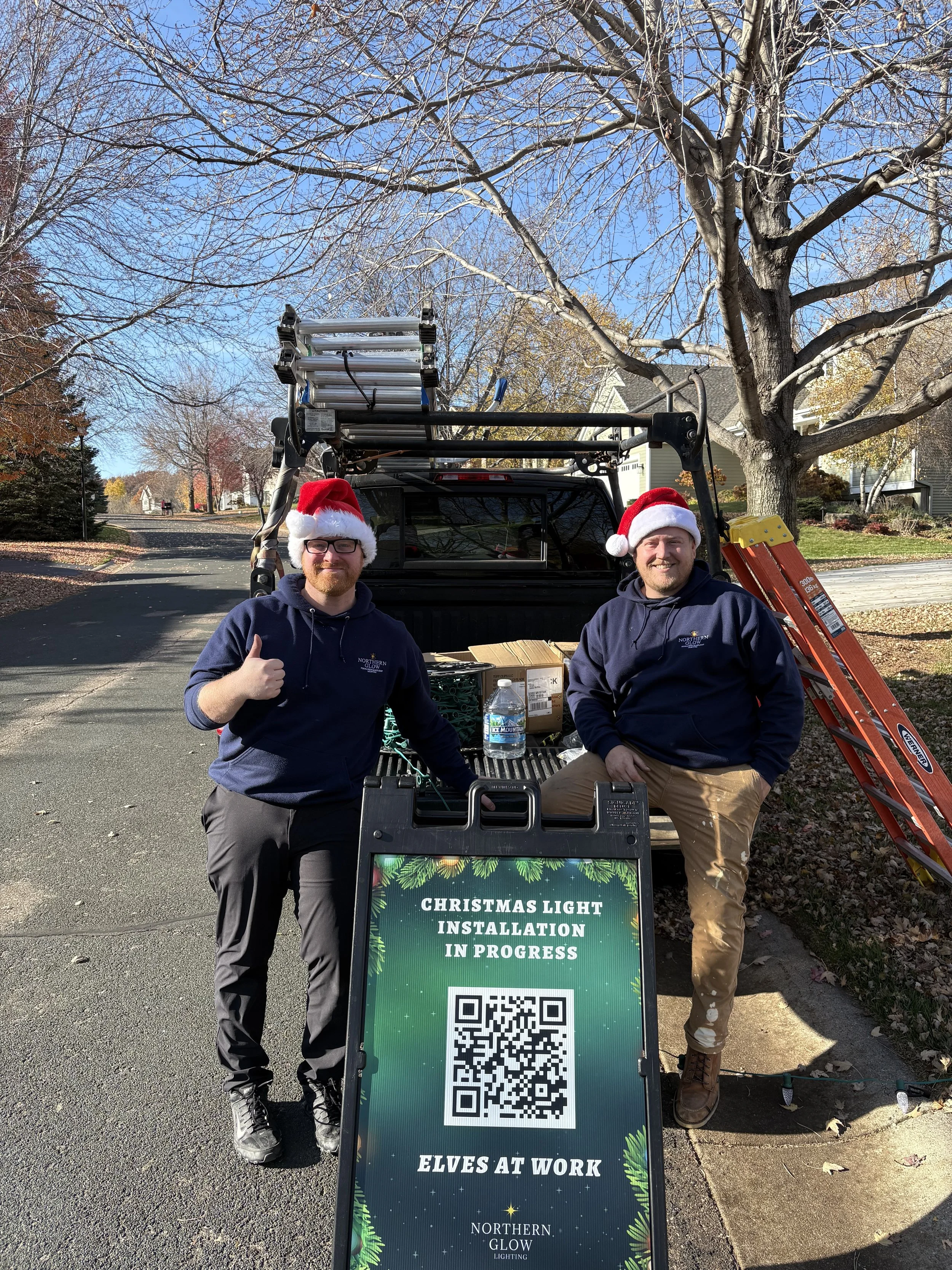 two men wearing santa hats and navy blue hoodies with Northern Glow Lighting logo on the chest - they are standing by a sign that reads "Christmas Light Installation in Progress"