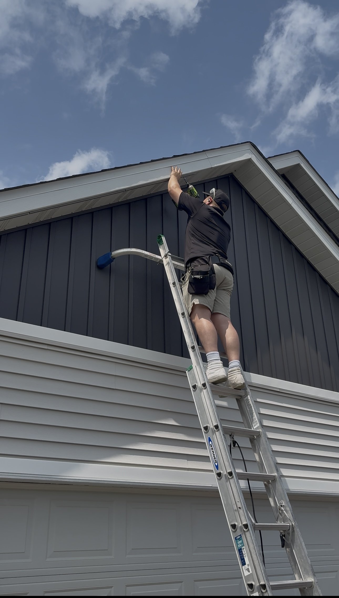 man in tan shorts and black shirt on tall ladder installing lights on a roof line above a garage