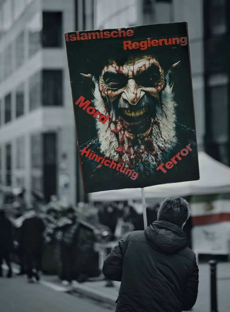 A person holding a protest sign featuring a graphic, monstrous face with horns, red and black coloring, and German words: 'Islamische Regierung', 'Mord', 'Hirntichtung', and 'Terror.'