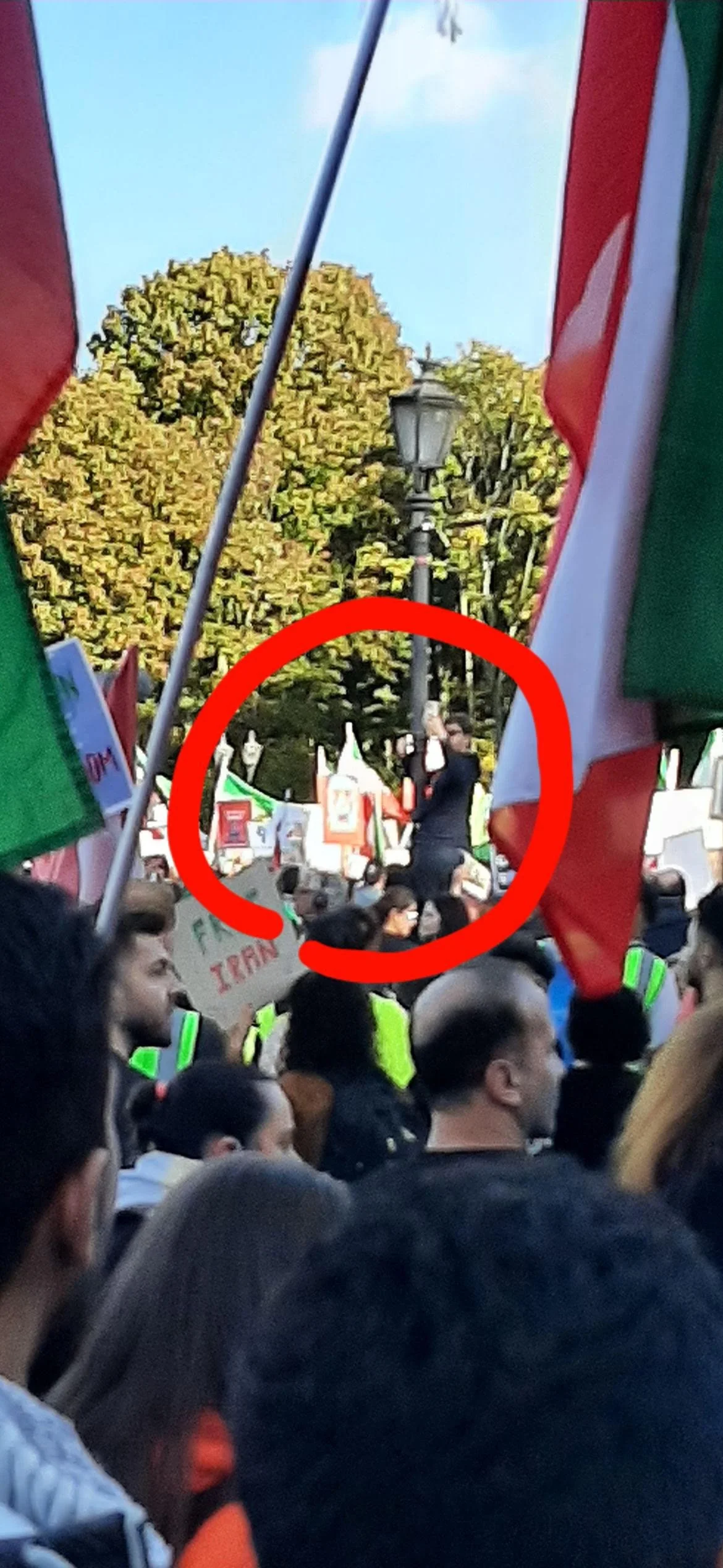 Crowd of people at a rally or protest, some wearing safety vests, with flags and banners. A person is standing on a platform or stage, speaking to the crowd, and is circled in red.
