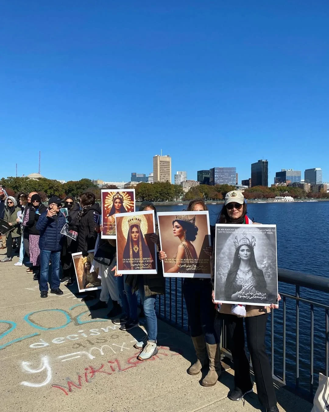 People holding posters with images of women, some wearing crowns and religious icons, standing by a waterfront with city buildings in the background.