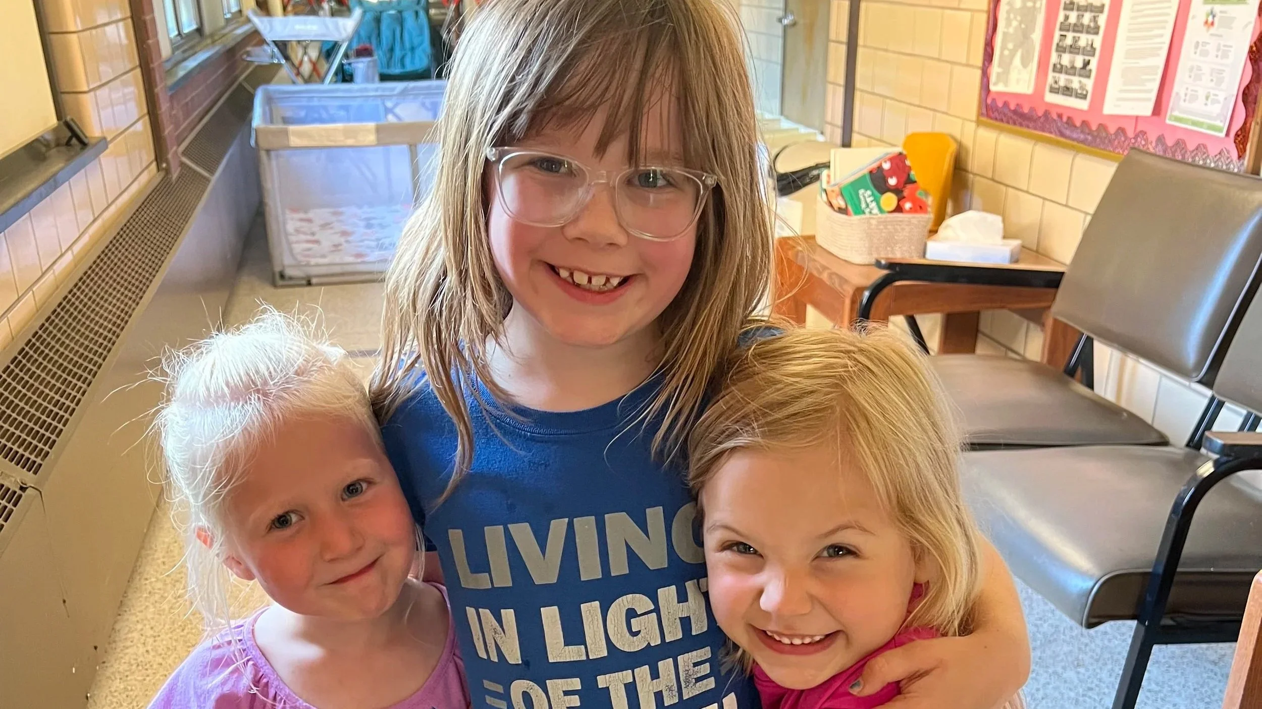 A smiling young girl with glasses and long hair, surrounded by three other children, in a school or community room with chairs and a pink bulletin board in the background.