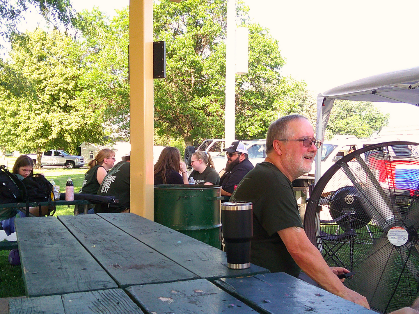 People sitting at a picnic table outdoors during daytime, with a man in the foreground near a large fan and several others in the background, surrounded by trees and parked cars.