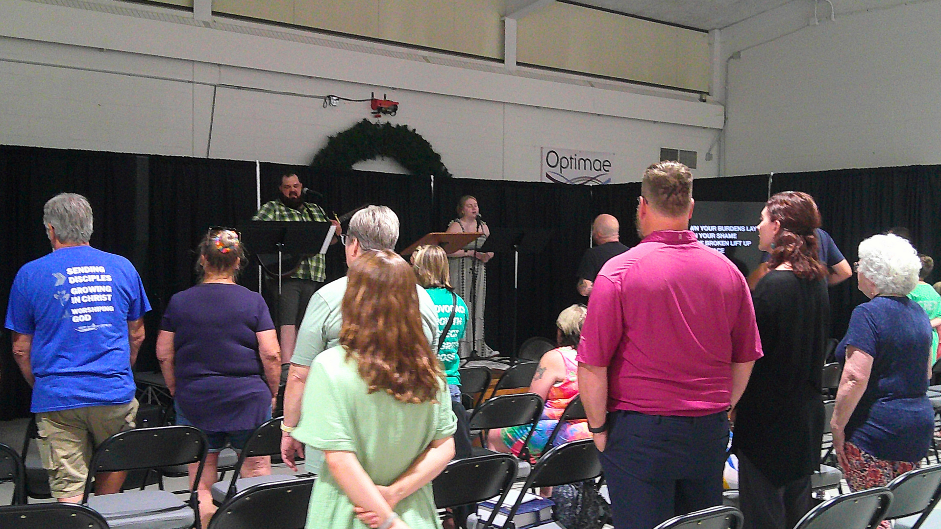 People attending a church service with musical worship. The congregation is standing, facing the stage where two musicians are performing. The stage has a black curtain backdrop, a Christmas wreath hangs on the wall, and a sign with the word 'Optima' is visible. The audience includes people of various ages, some with gray hair, and someone wearing a blue T-shirt with a religious message.