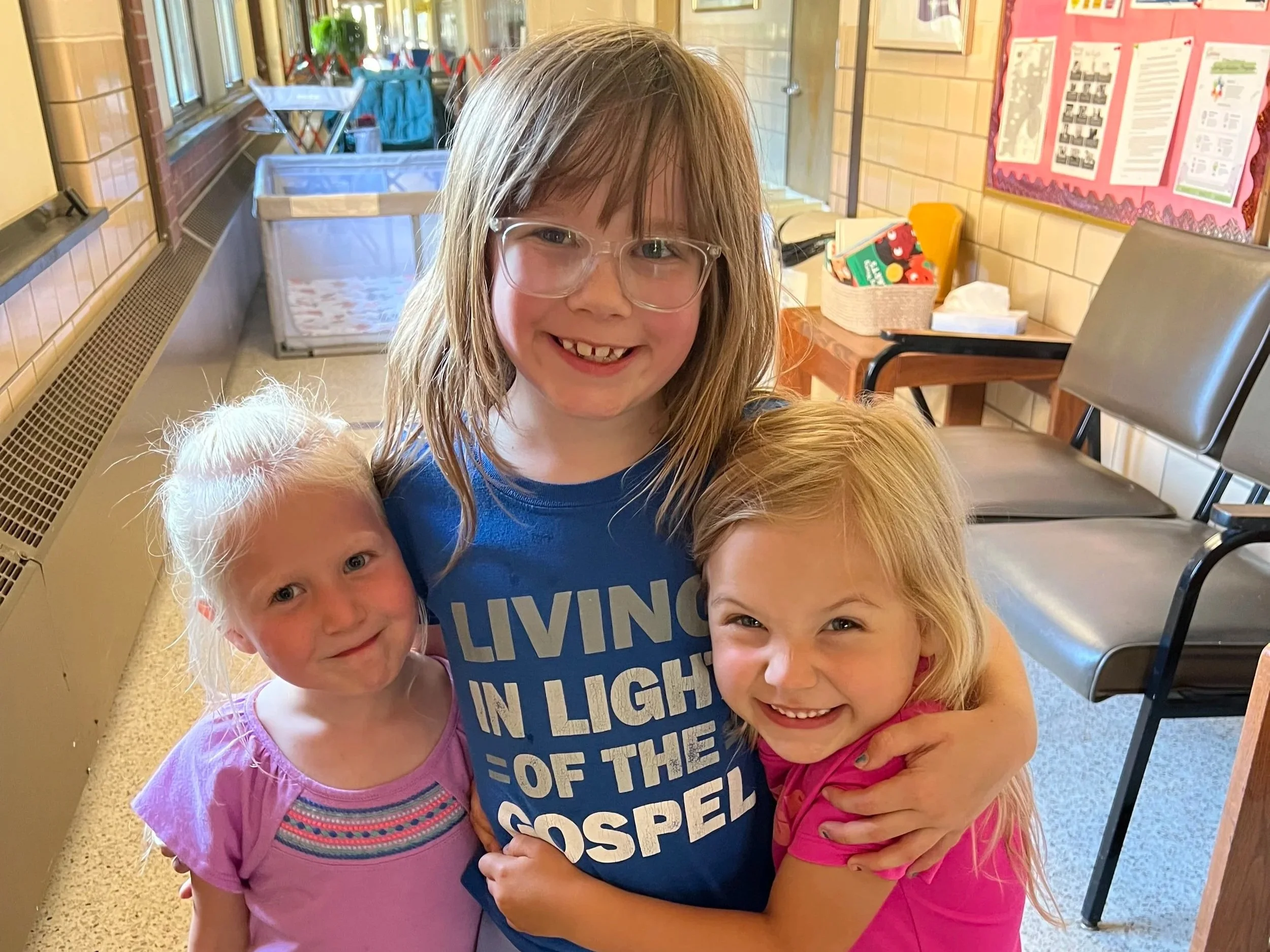 A smiling girl with glasses and long hair hugging a group of younger children, in a school or daycare setting with chairs and a bulletin board in the background.