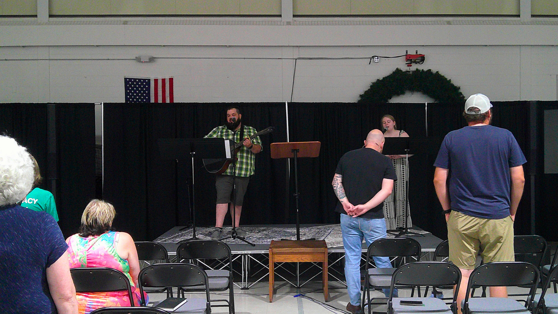 People attending a small indoor musical performance with two singers and a guitarist on stage. Audience members are seated and standing, watching the performers in front of a black curtain with an American flag and holiday wreaths on the wall.