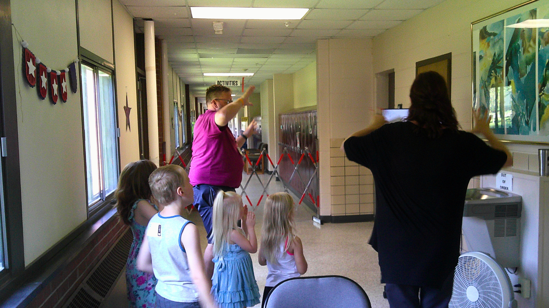 A group of young children and two adults in a hallway of a school, participating in an activity. One adult is directing the children, while the other is taking a photo or video. The hallway has windows and decorations, including a sign that says 'ACTIVITIES'.