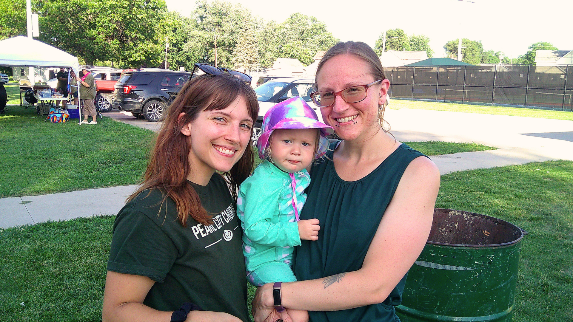 Three women, one holding a young girl, smiling outdoors at a park or event area with vehicles and a tent in the background.