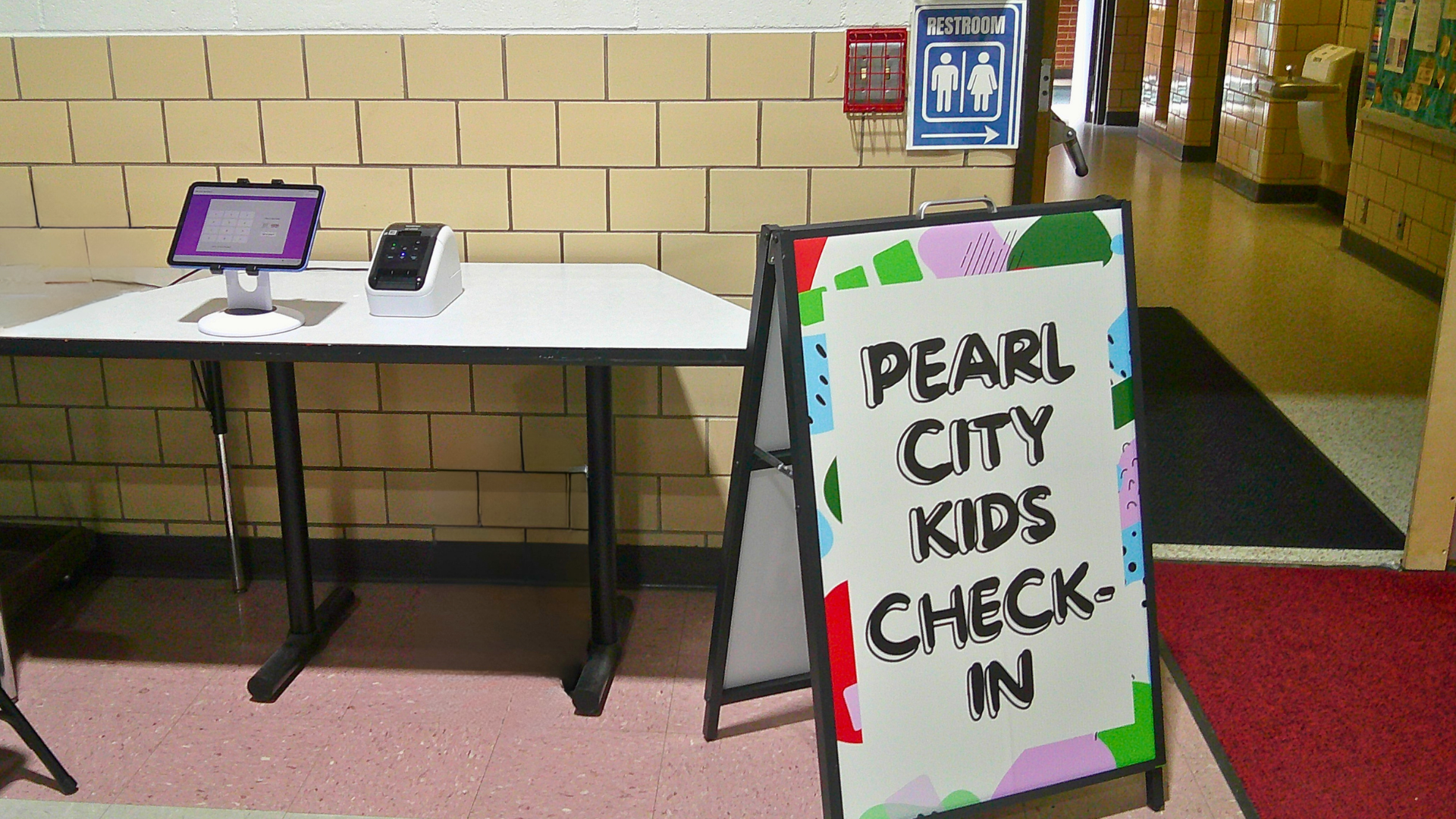 A table set up outside a school with a sign that reads "Pearl City Kids Check-In". There is a tablet and a device on the table. A restroom sign is visible on the wall behind the table.