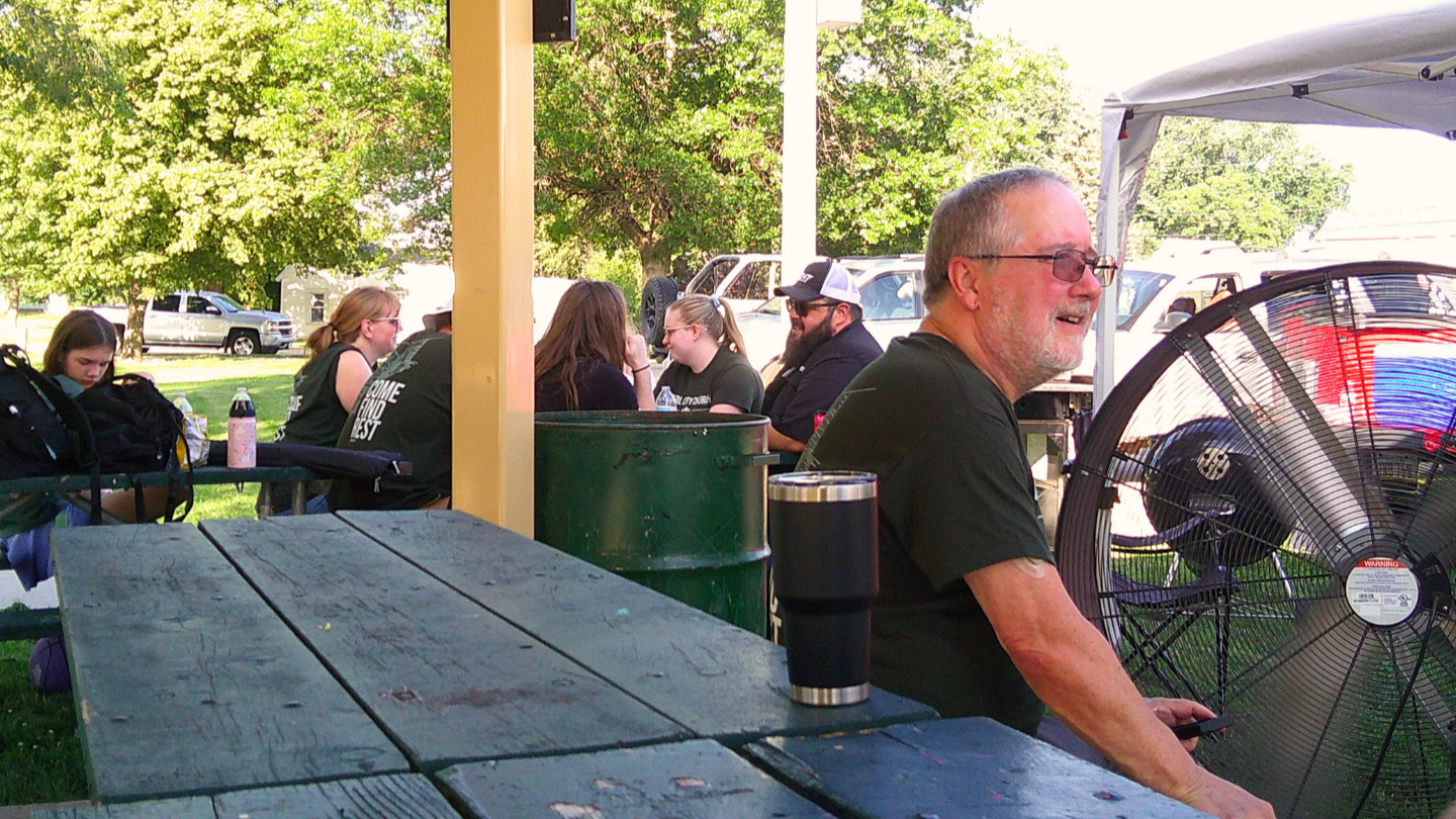 People sitting at a picnic table outdoors, some engaged in conversation, with a man in sunglasses smiling nearby and a large fan in the background.