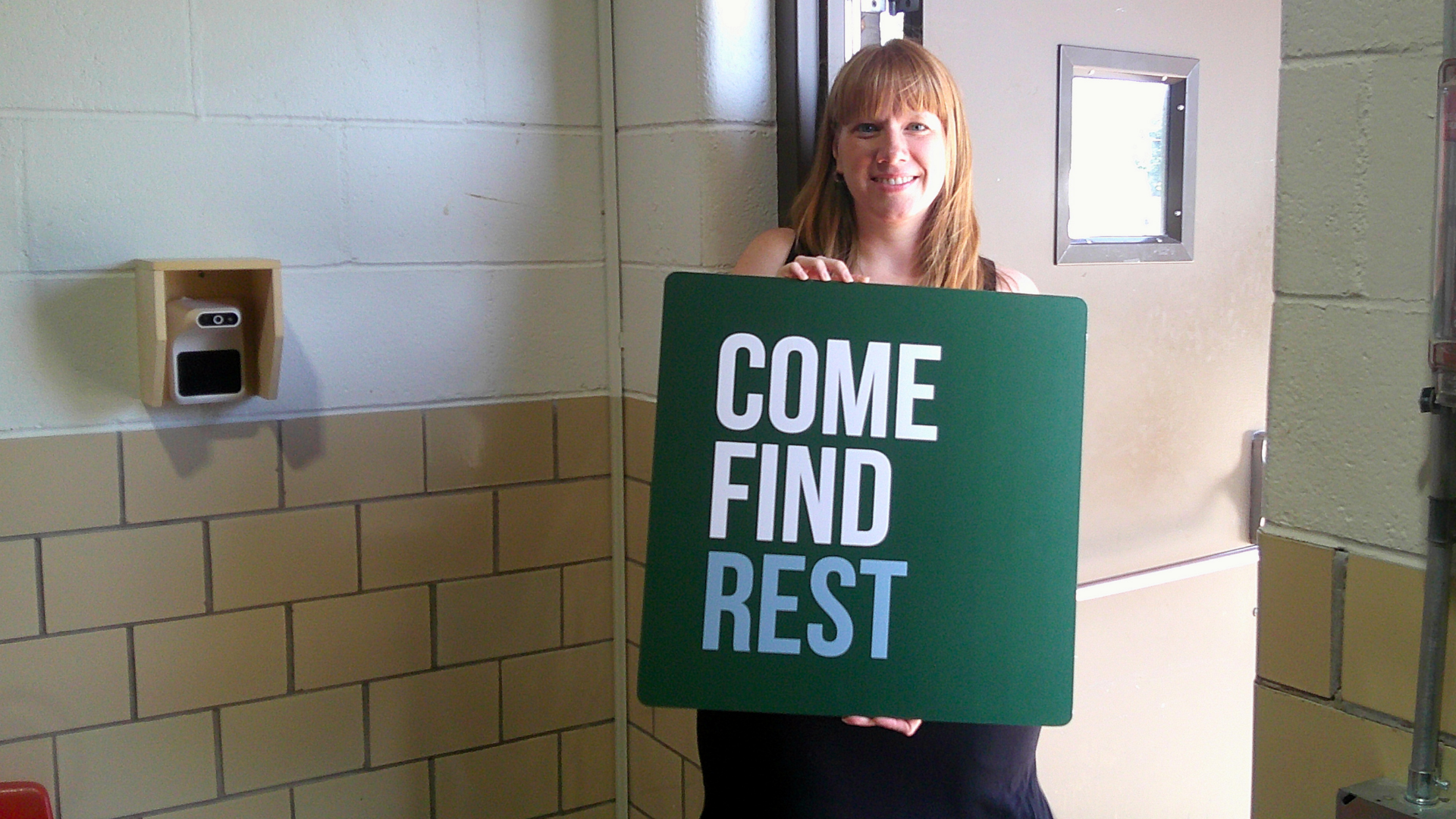 A smiling woman with red hair holding a green sign that reads "COME FIND REST" inside a building with beige brick walls and a small window.