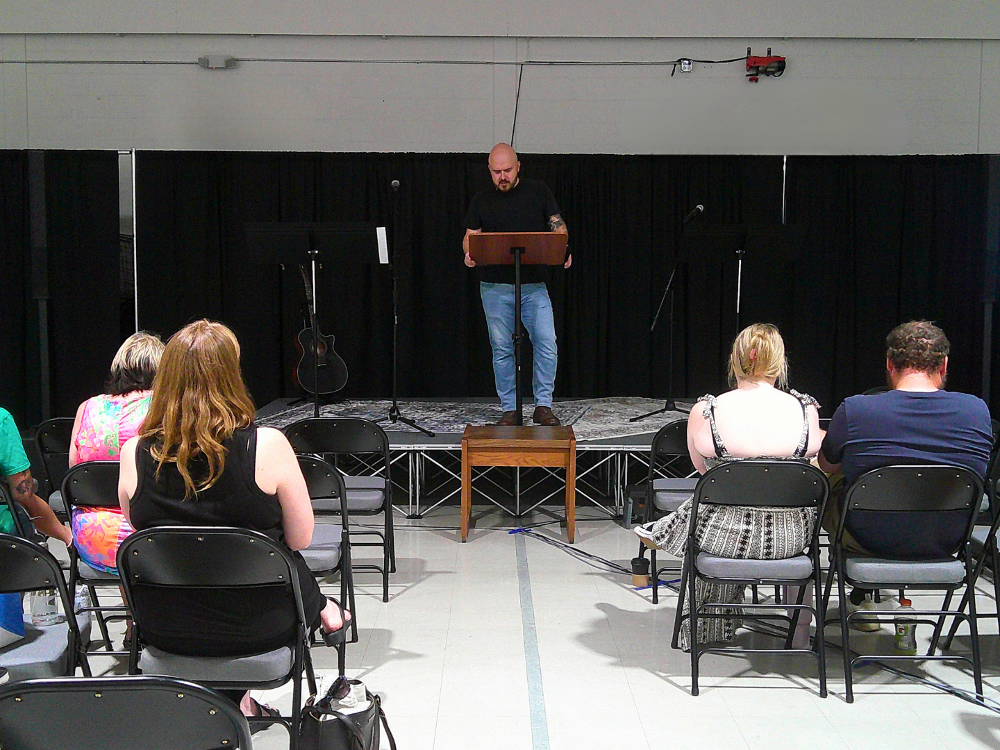 A small indoor stage with a man speaking at a podium, surrounded by musical instruments including guitars and microphones, with an audience seated in rows watching him.