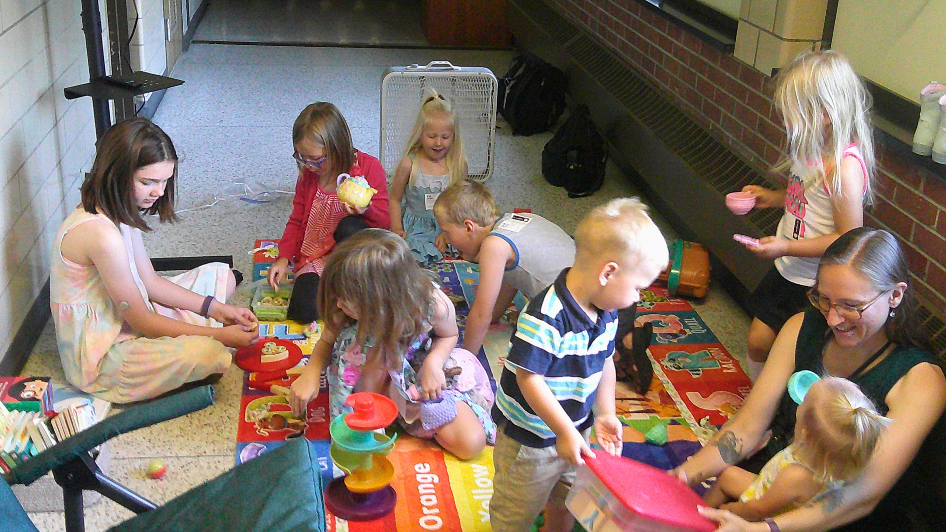 Children and adults sitting on a colorful mat playing with toys in a hallway.