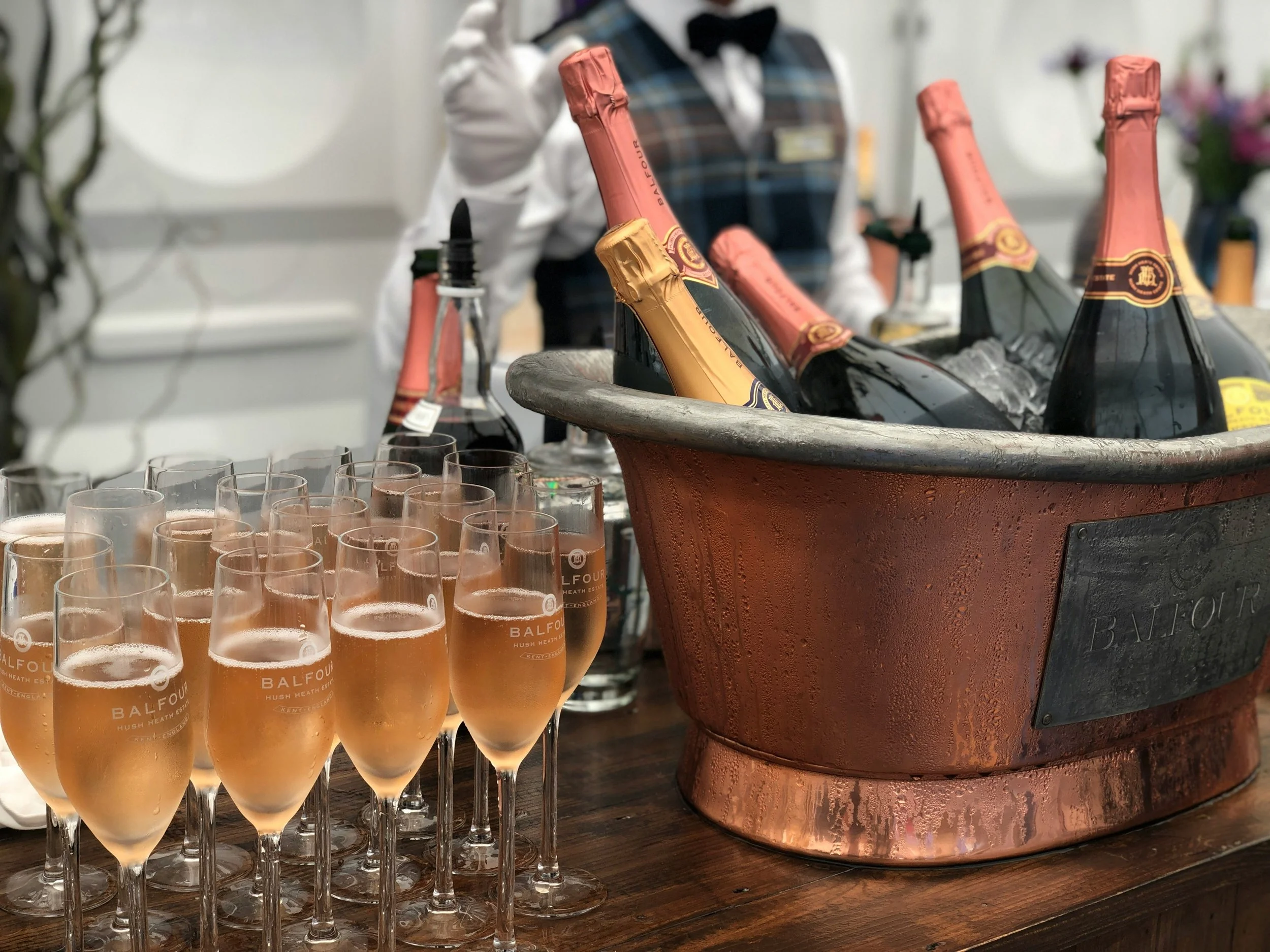 A wine cooler filled with bottles of sparkling wine, alongside around twenty glasses of pink sparkling wine