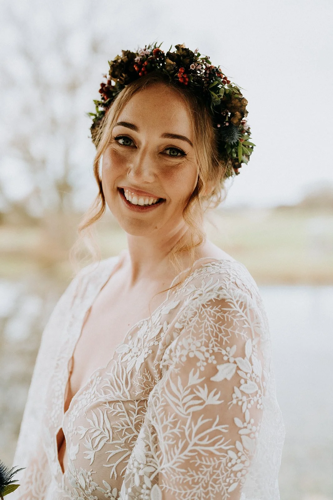 Kimberley Jones, a smiling woman with a floral crown stands outdoors near a body of water. She wears a lace wedding dress with floral embroidery.