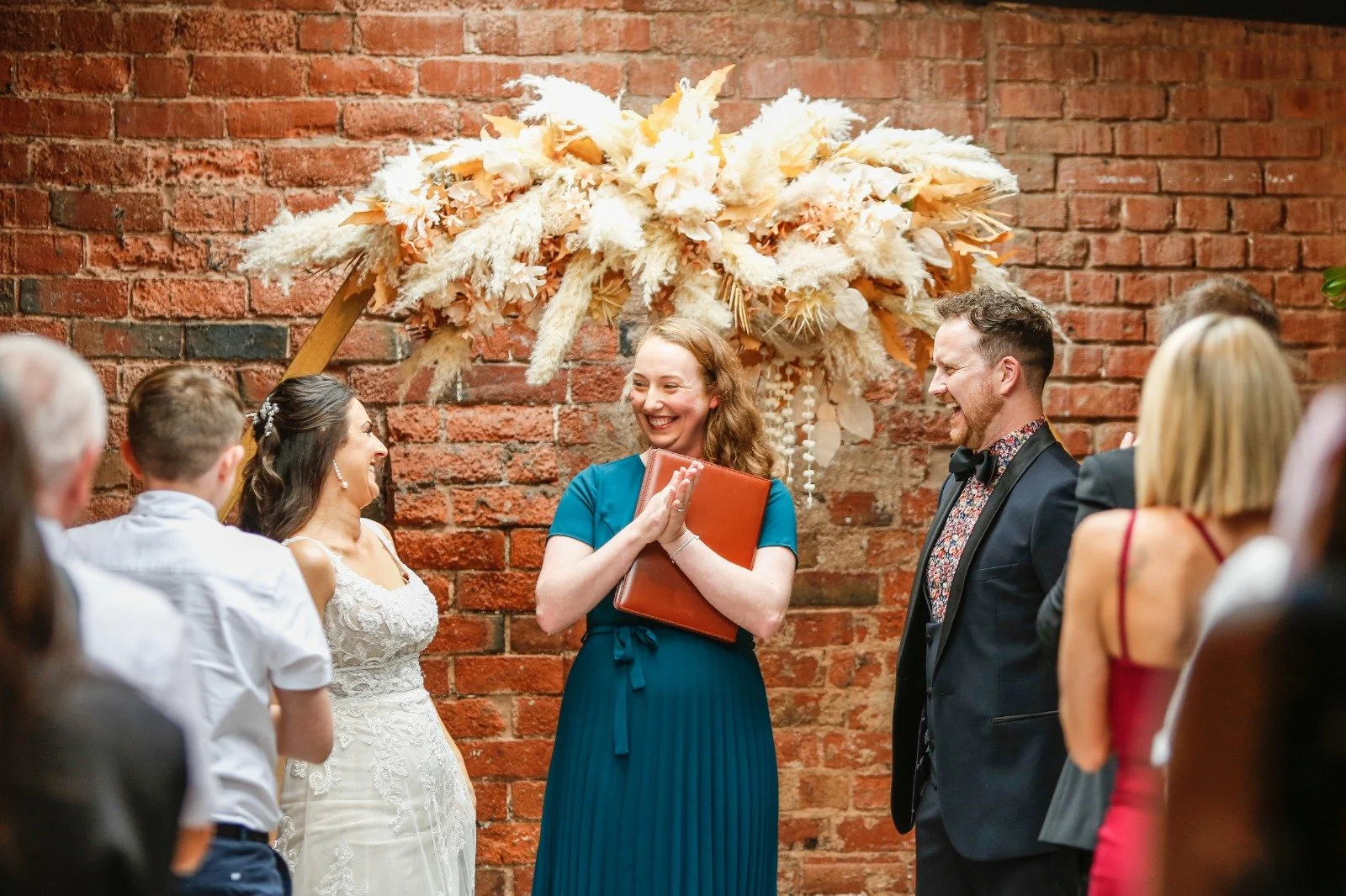 A wedding ceremony with a bride and groom standing facing Kimberley Jones, with friends and family gathered around, against a red brick wall with a large dried flower arrangement overhead.