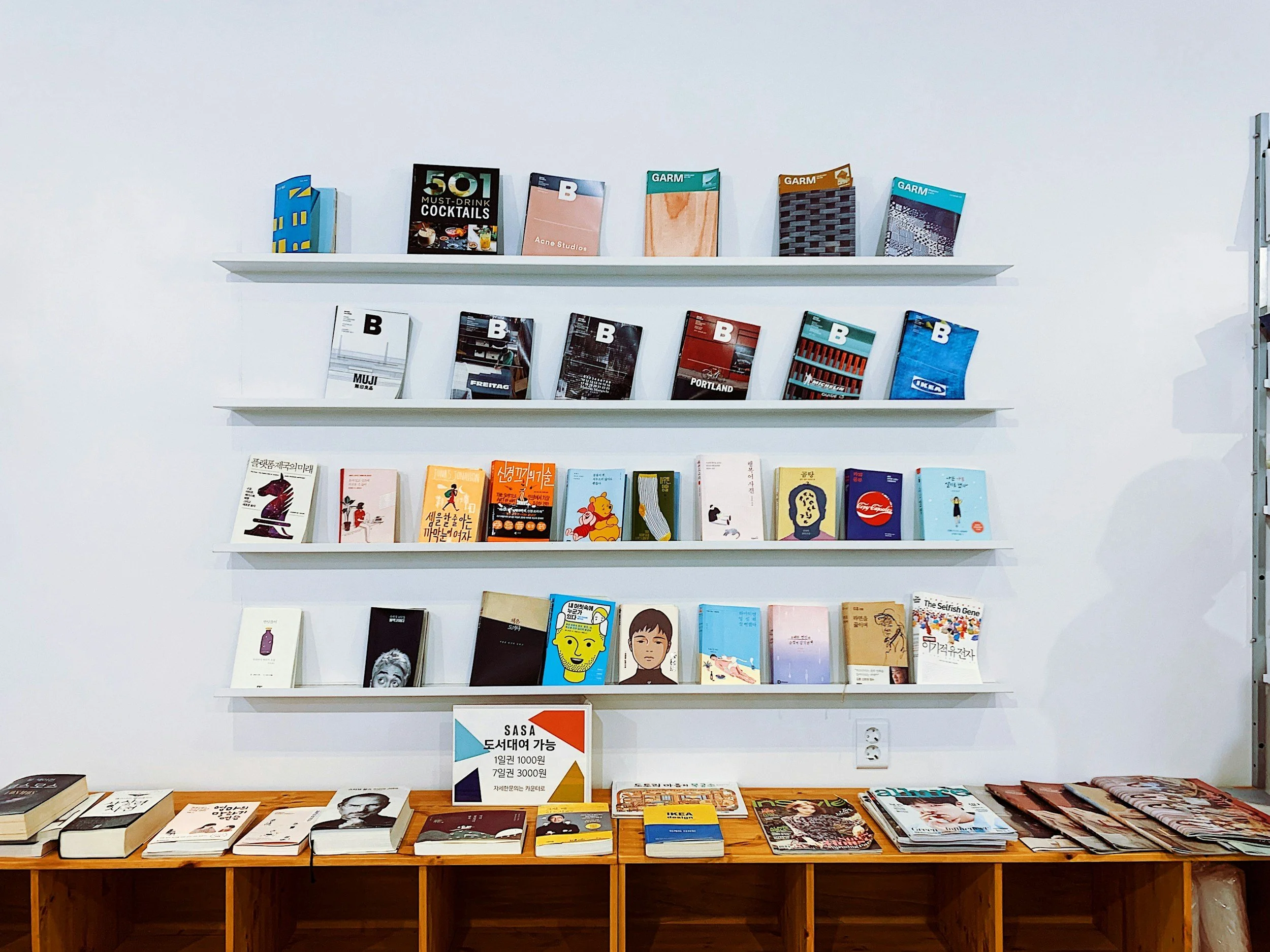 Books displayed on four white shelves with various titles and covers, and additional books on a wooden table below, against a plain white wall.